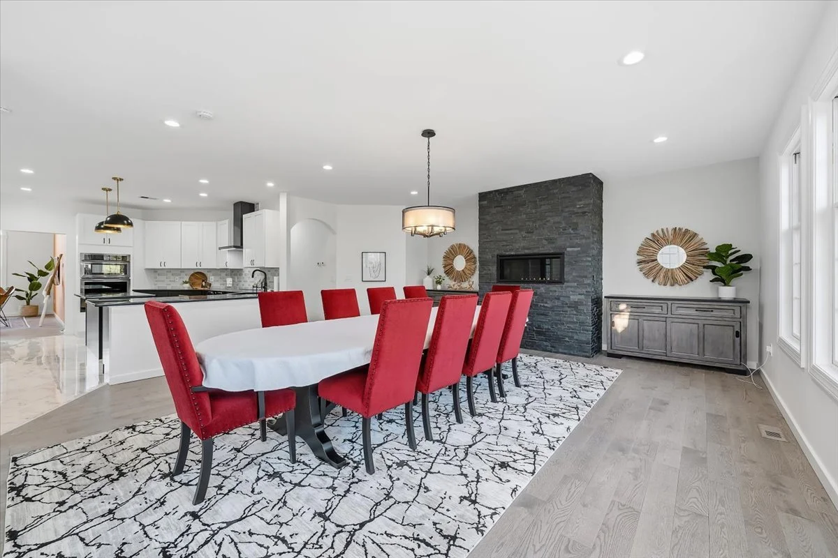 Modern dining room with a long white table and red upholstered chairs, black and white patterned rug, dark stone fireplace, gray cabinet, and minimalist decor.
