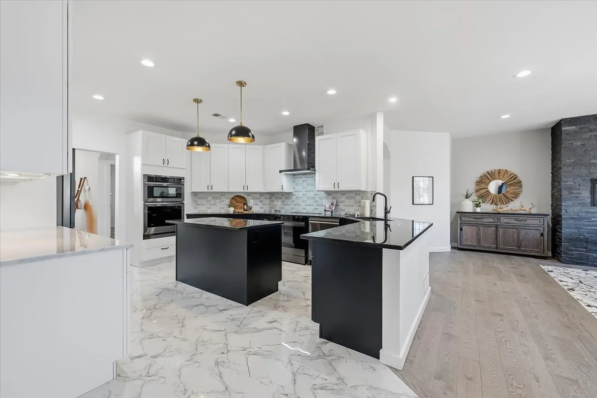 Modern kitchen with white cabinets, black countertops, and a central island. The kitchen features double wall ovens, a stove with a range hood, and pendant lights. The floor is a marble pattern, and there's a small adjacent dining or living area with