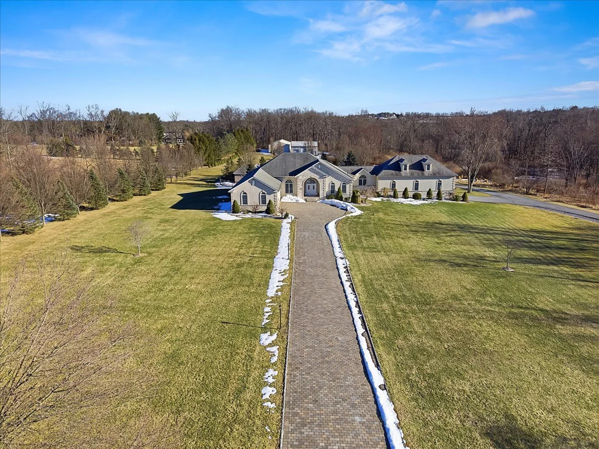 A large house with a gray roof and light-colored exterior, set on a spacious plot with a long brick driveway, surrounded by a lawn with patches of snow, and trees in the background under a blue sky.
