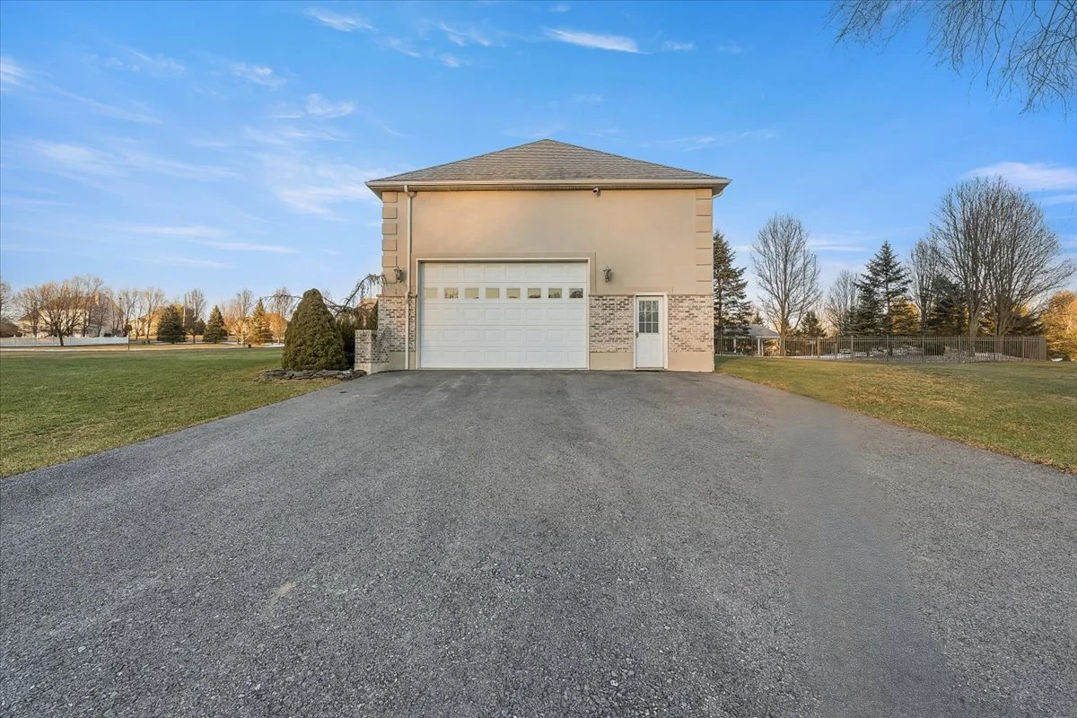 A two-story beige house with a white garage door and a small white side door, surrounded by open grassy area and trees, with a paved driveway leading up to the garage.