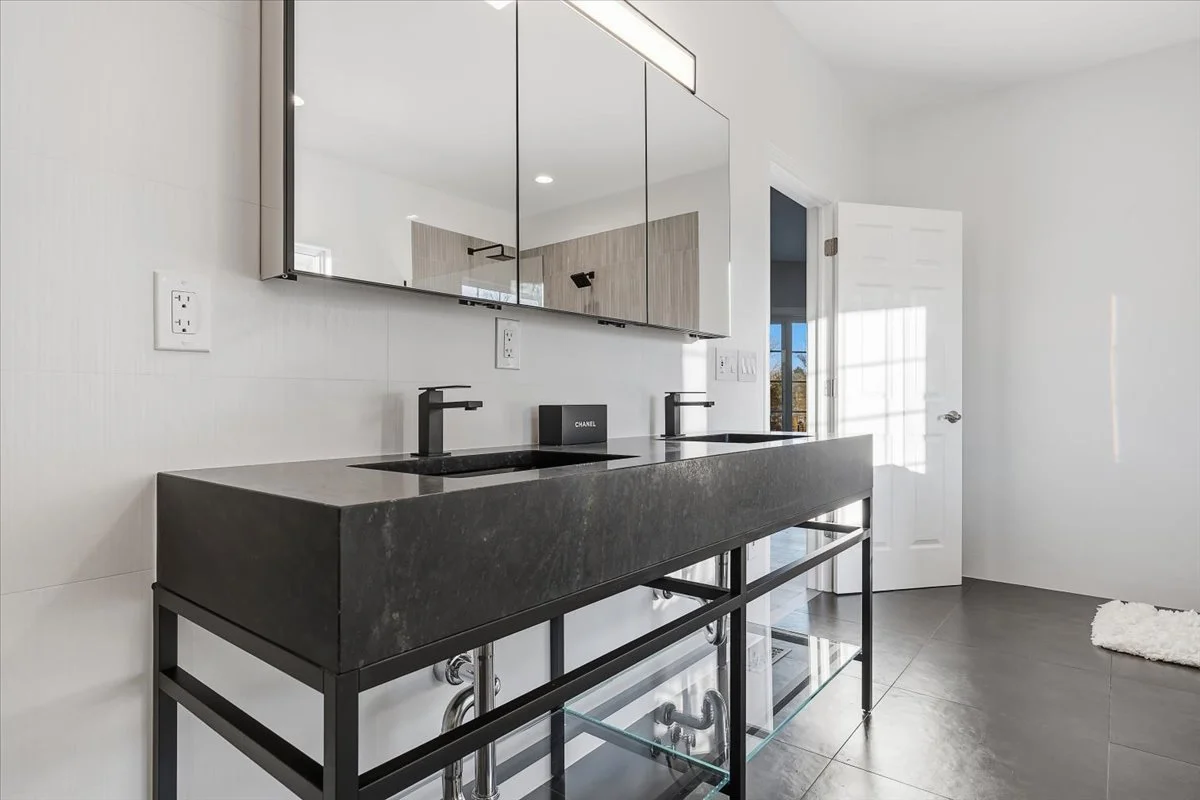 Modern bathroom with double black sinks, large mirror with cabinet, white walls, dark gray floor, open door with sunlight, white fluffy rug.