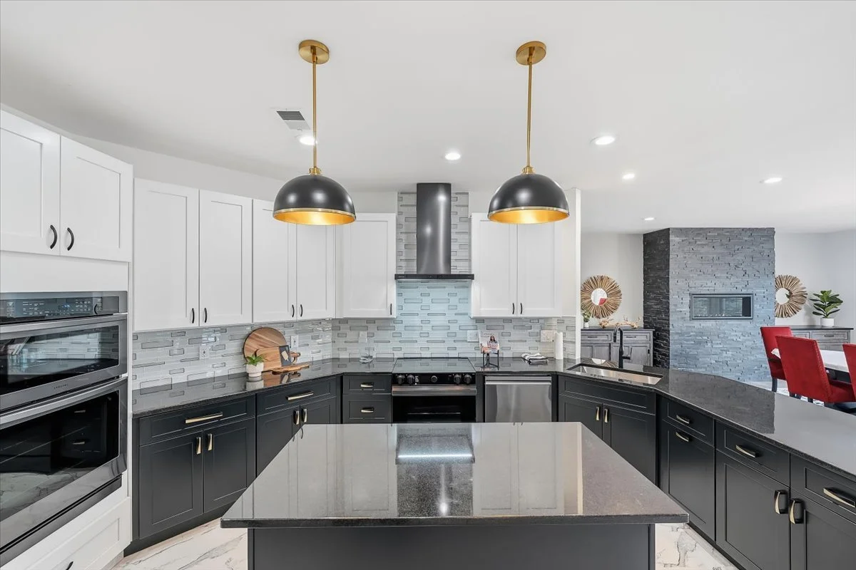 Modern kitchen with black and white cabinets, a gray tile backsplash, black countertops, two black pendant lights with gold interiors, and stainless steel appliances.