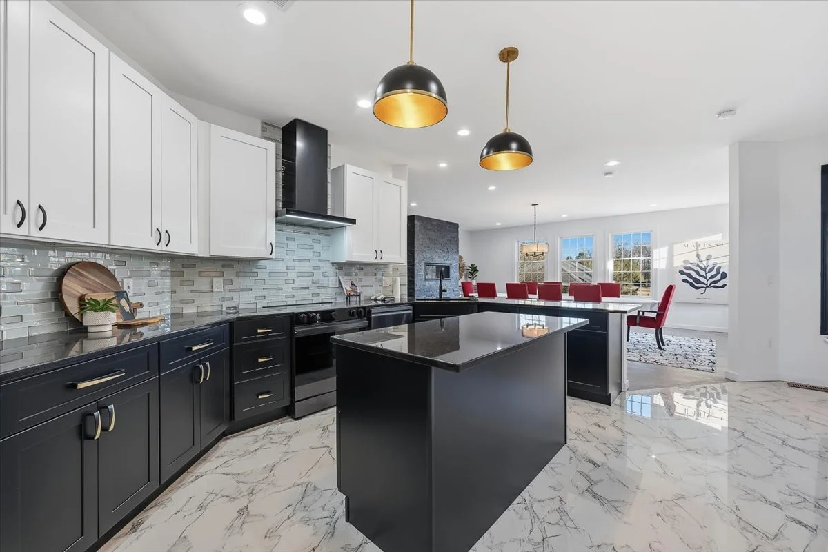 Modern kitchen with white upper cabinets, black lower cabinets, and a black kitchen island with a glossy black countertop. The floor is white marble with gray veining. Hanging black and gold pendant lights are above the island. In the background, the