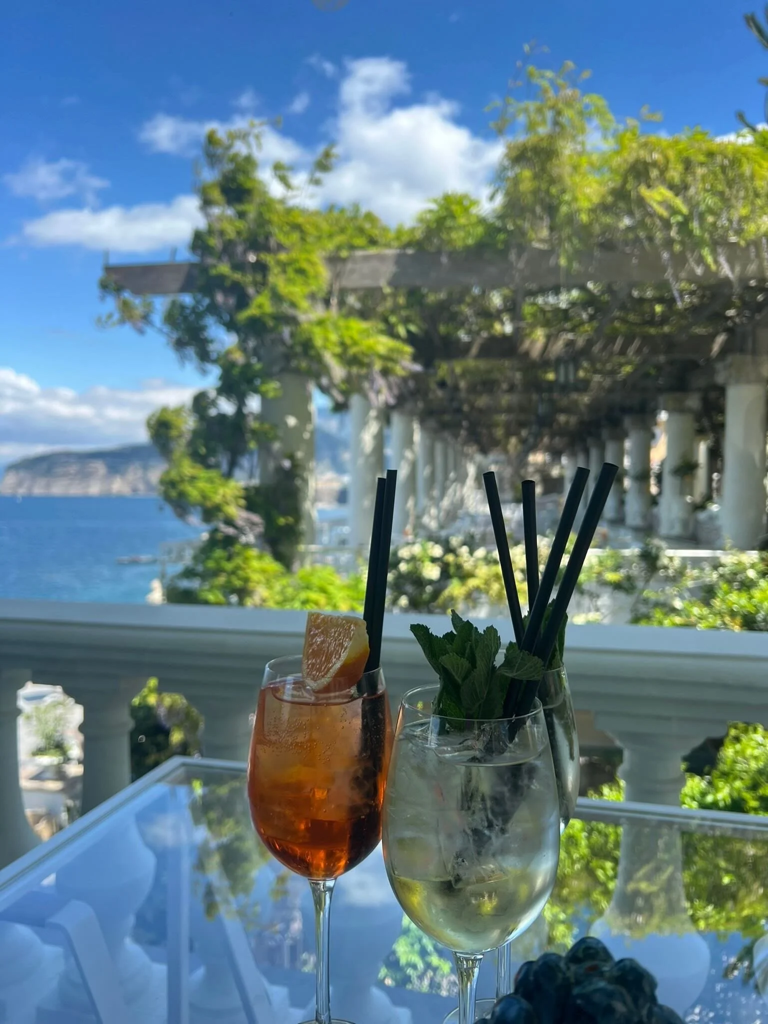 Three drinks on a glass table with a scenic seaside view behind - Travel - KPJ Collective