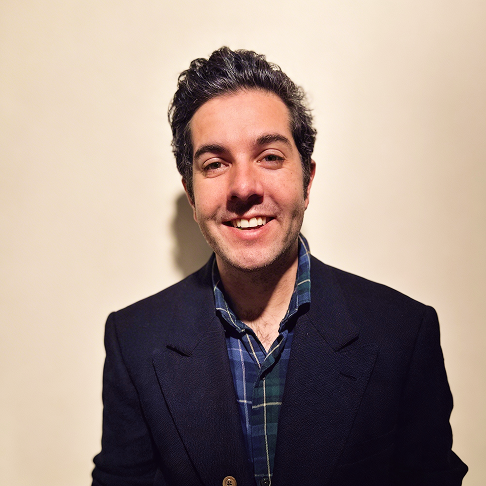 A man with dark, curly hair smiling, dressed in a dark blazer and plaid shirt, standing against a plain light-colored wall.