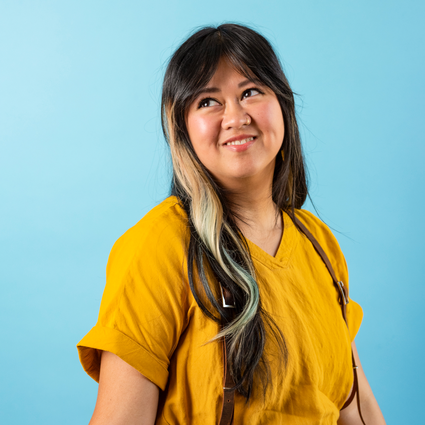 A woman with long, dark hair with blonde and turquoise streaks, wearing a mustard yellow shirt, smiling and looking to the side against a light blue background.