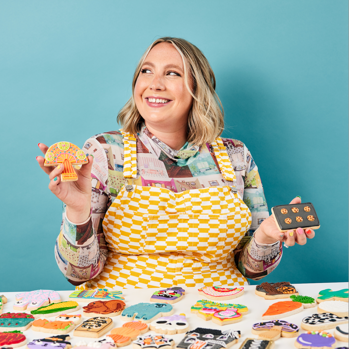 A woman with shoulder-length blonde hair, smiling, holding decorated cookies and a tray of small cookies, standing behind a table filled with various decorated cookies against a blue background.