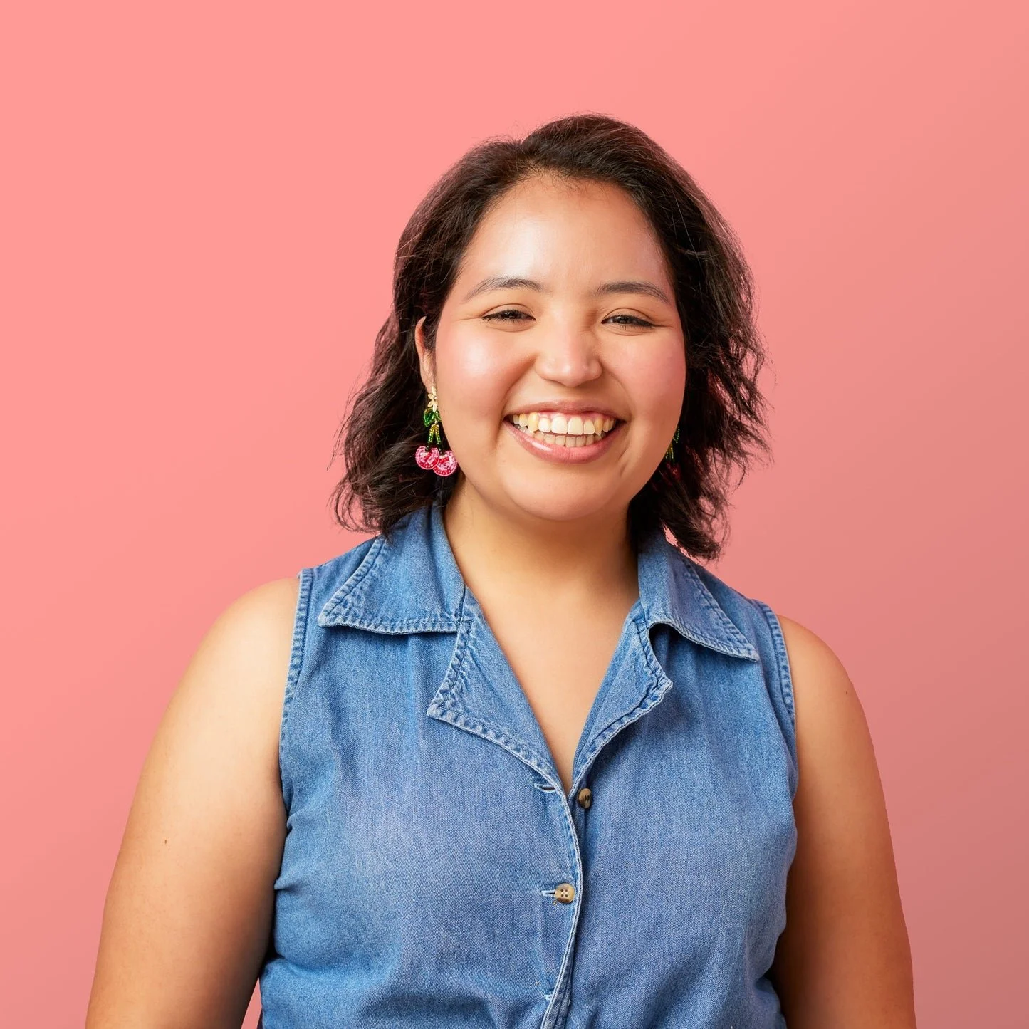 A smiling woman with curly dark hair wearing a sleeveless denim shirt and cherry earrings standing against a pink background.