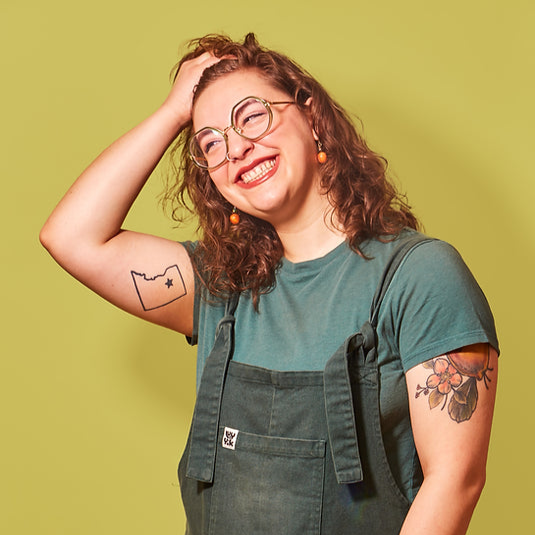 Young woman with curly hair, glasses, and tattoos, smiling with her hand on her head against a green background.
