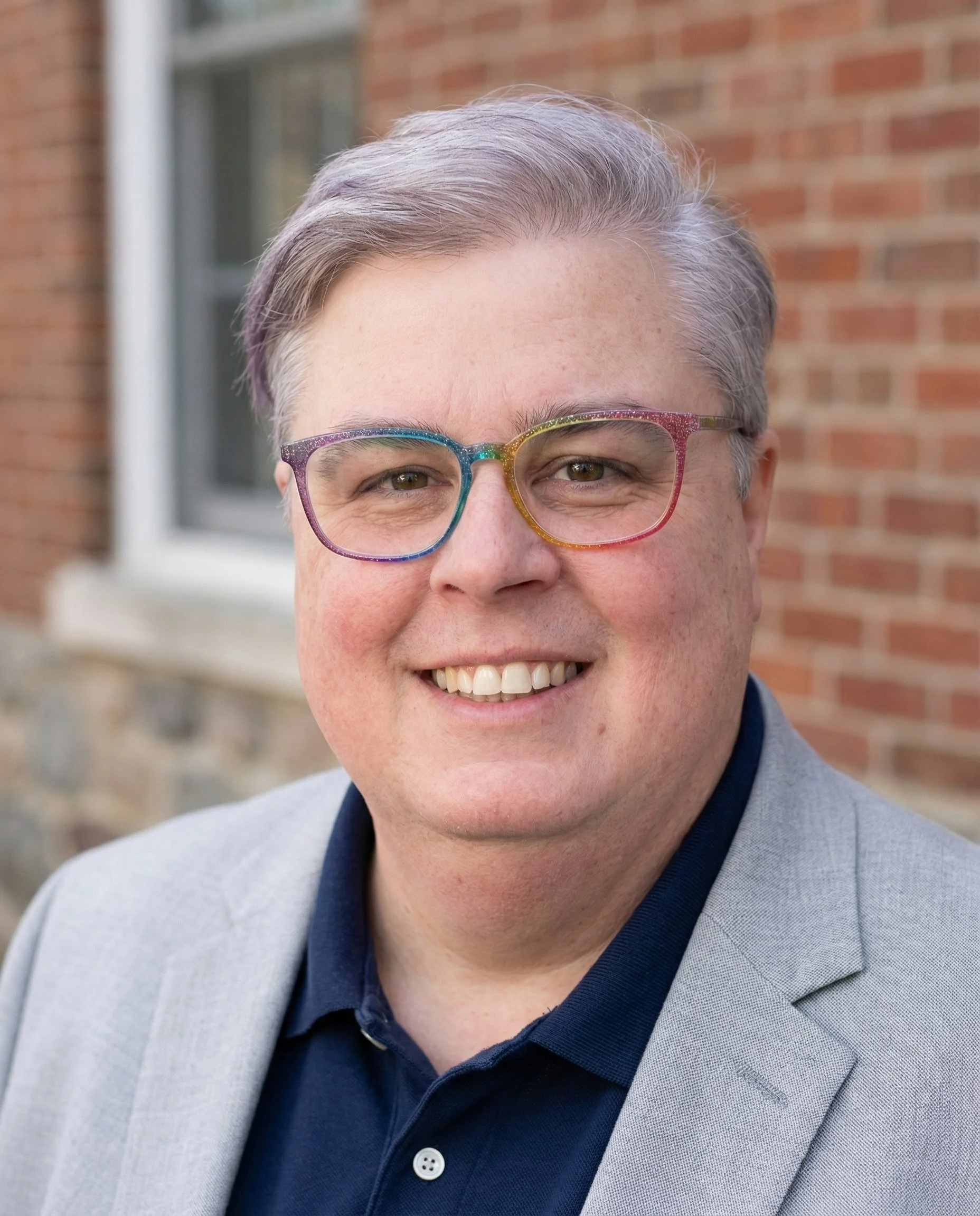 Headshot of a smiling person with short gray hair wearing colorful glasses, a dark shirt, and a light blazer, standing outdoors in front of a brick building.