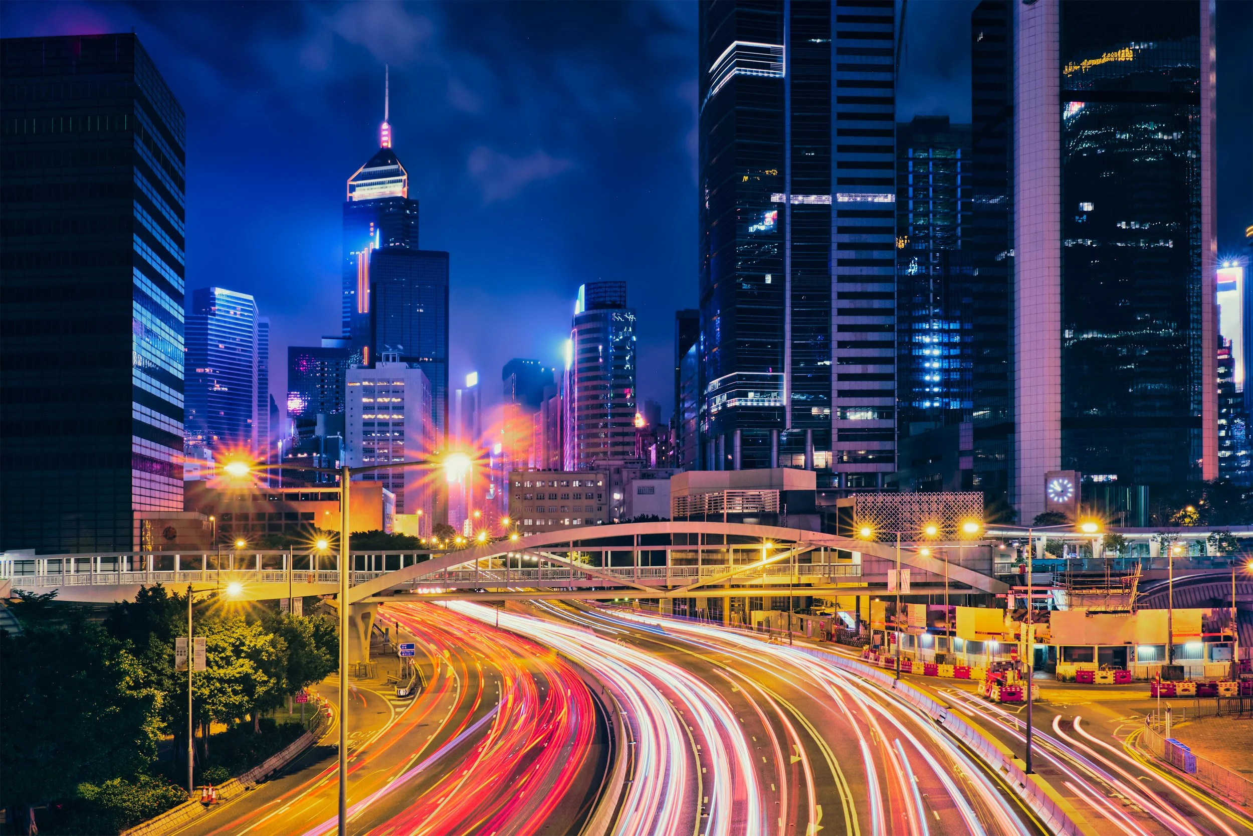 Nighttime cityscape with tall skyscrapers, busy highway with light trails from moving vehicles, and illuminated buildings in a downtown area.