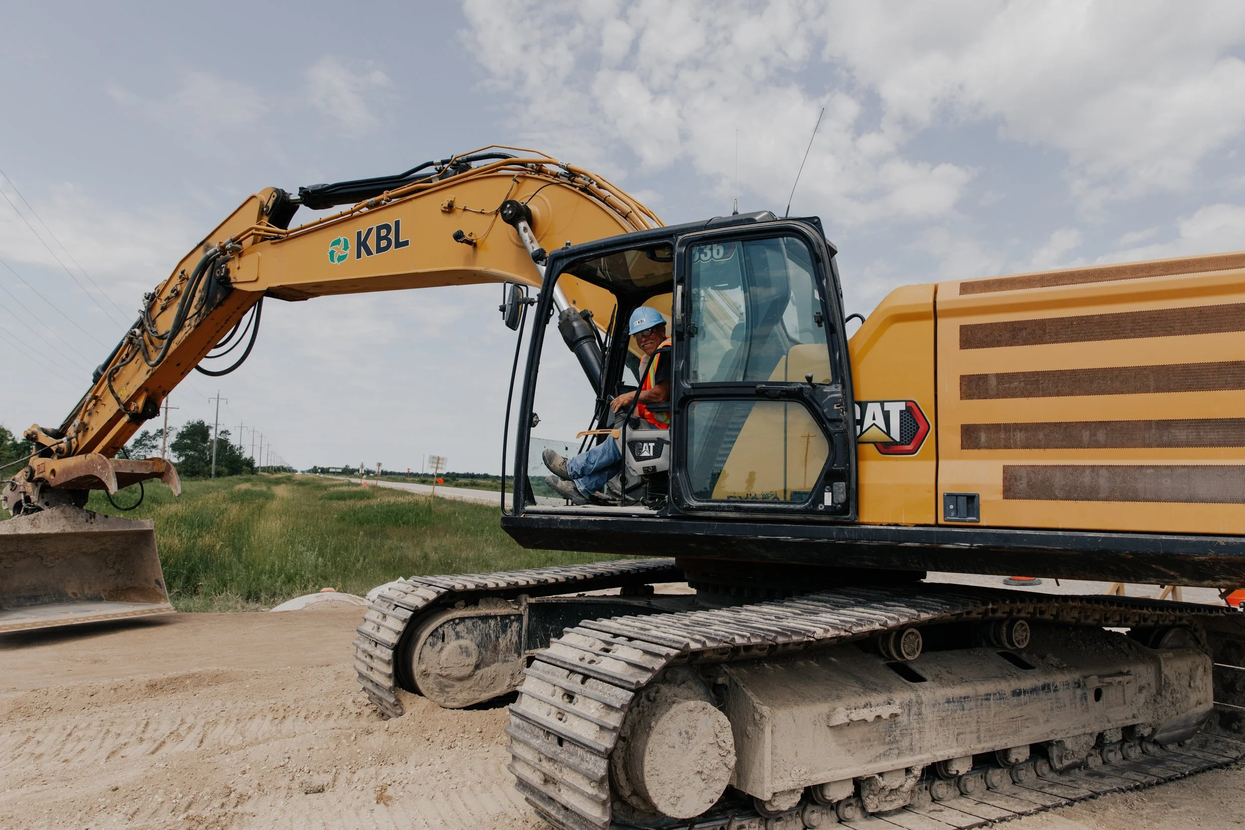 Construction worker operating a yellow CAT excavator on a construction site with dirt and a partly cloudy sky.
