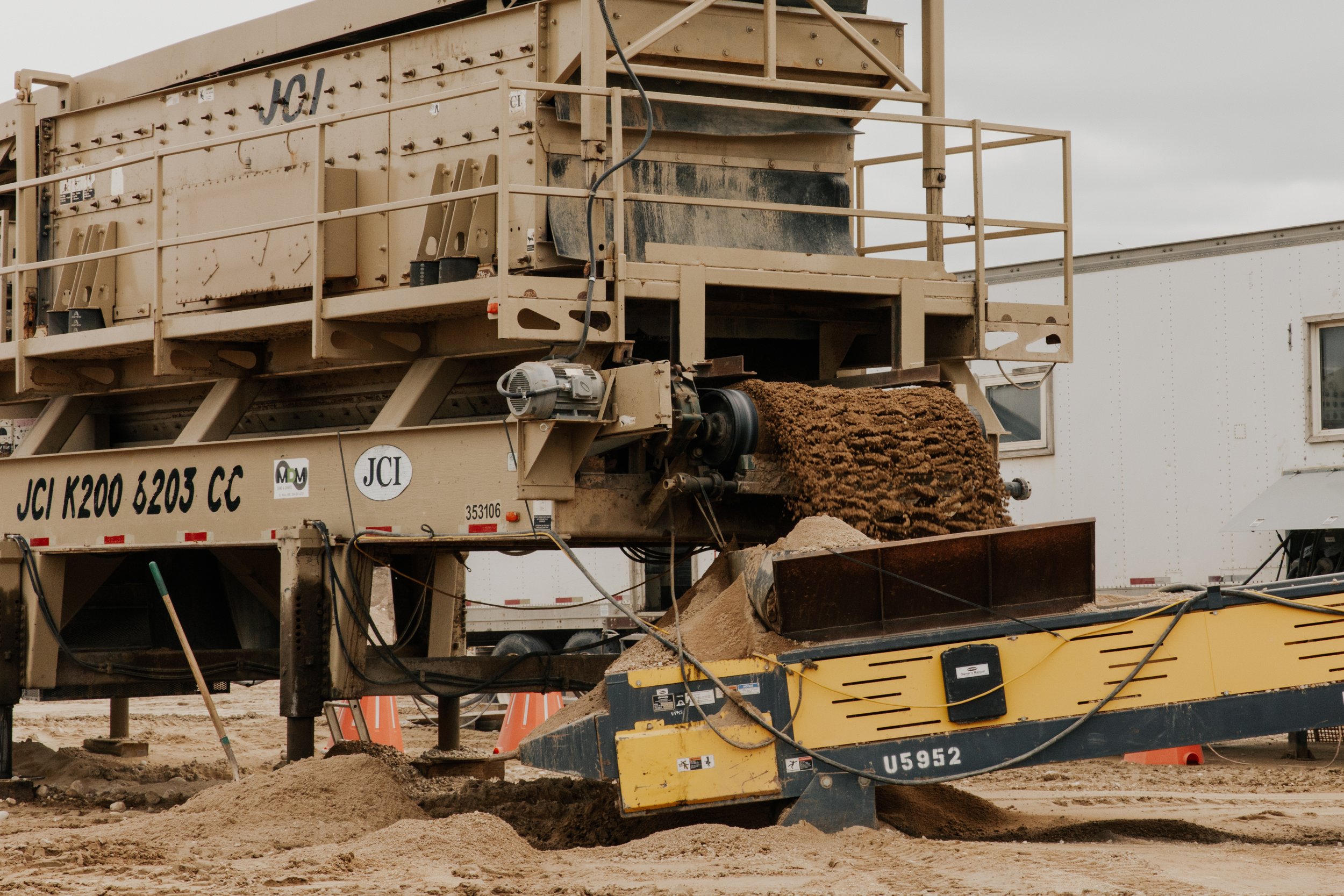 Heavy construction equipment on a dirt surface, including a large yellow machine and a beige asphalt paving machine, with dirt and construction cones in the background.