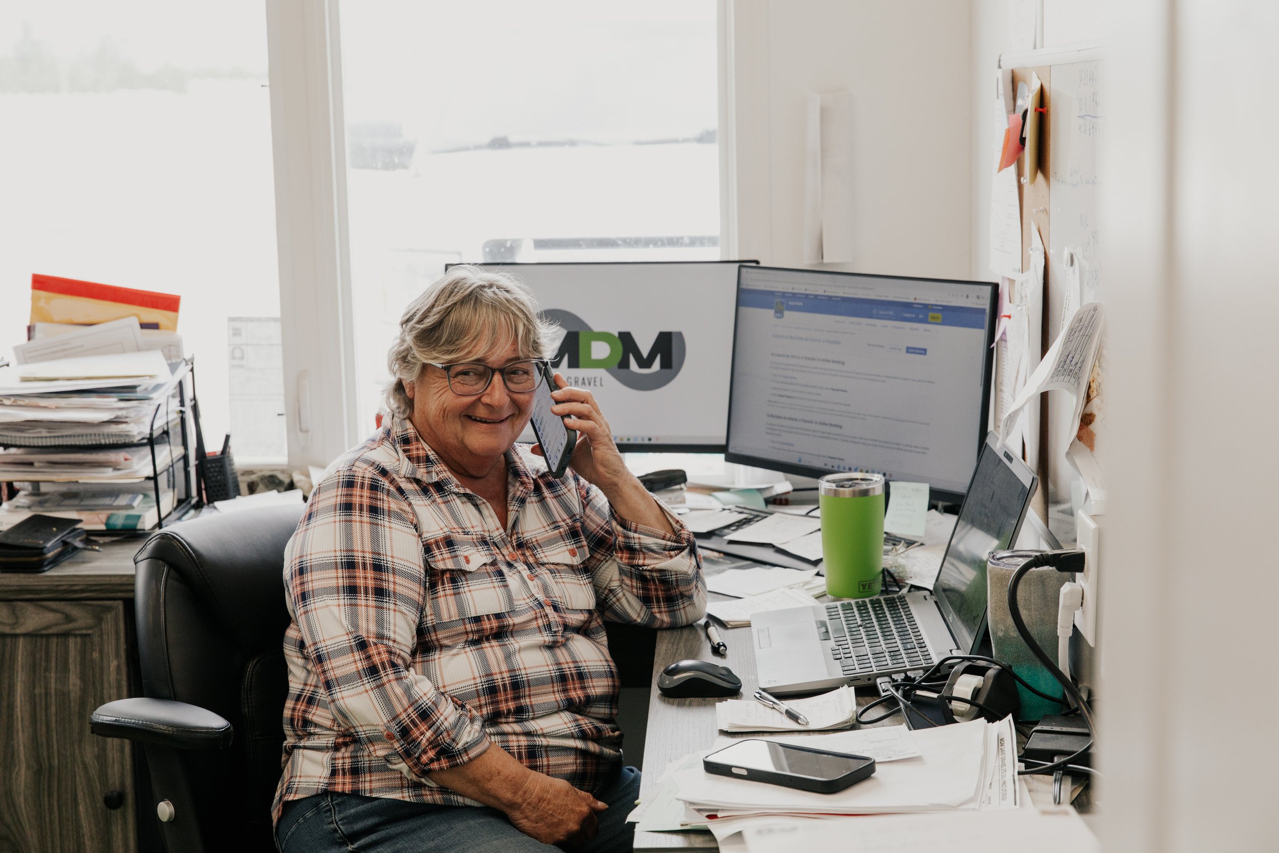 An older woman with gray hair and glasses, wearing a plaid shirt, smiling while talking on a cell phone in her cluttered office space with multiple computer monitors, papers, and office supplies.