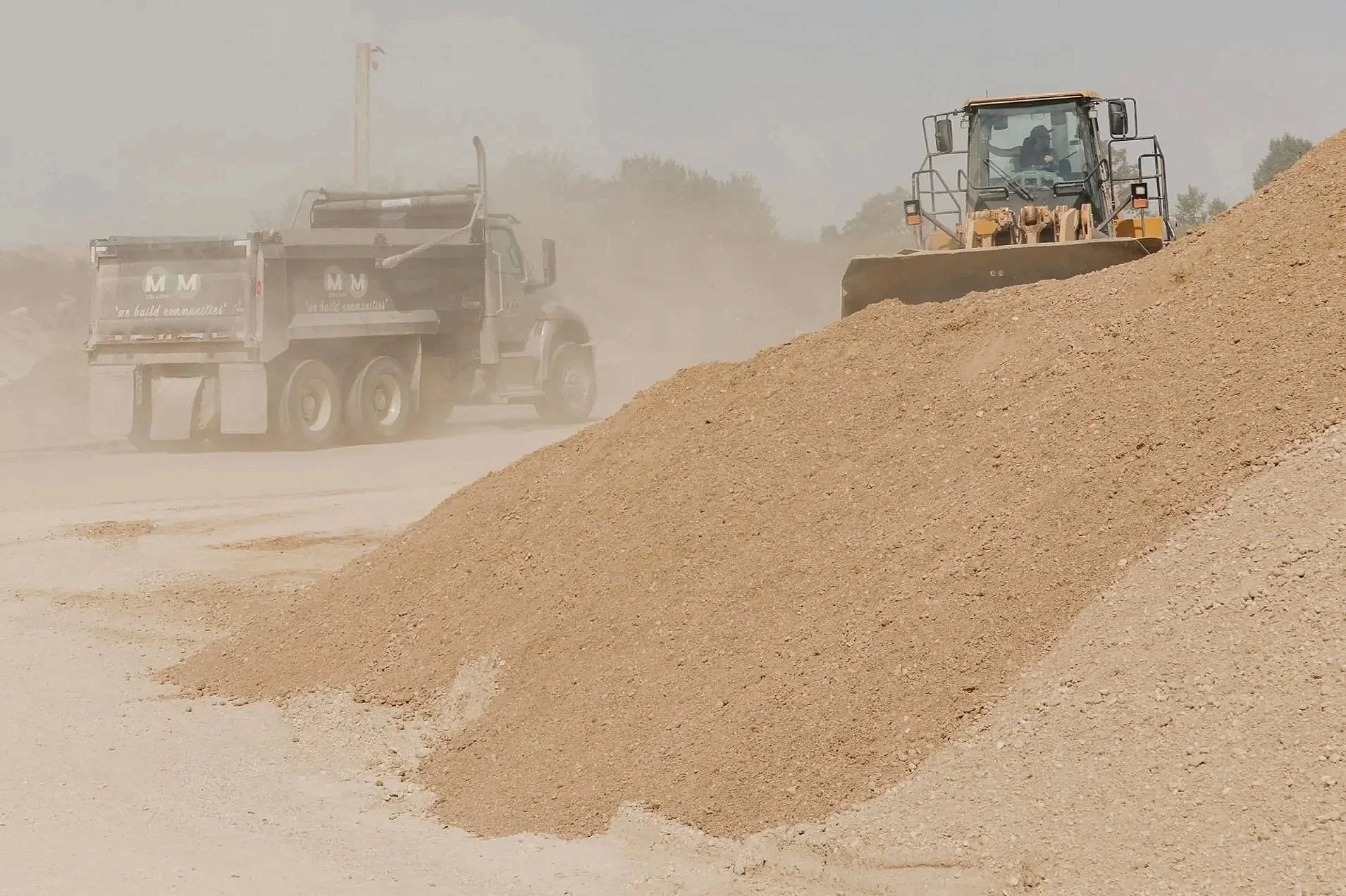 A construction site with a bulldozer pushing dirt and a dump truck driving behind, both working amidst a dusty environment.