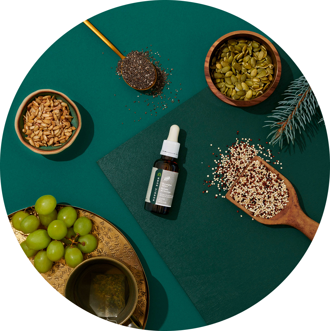 A flat lay of a wellness setup with various natural and herbal items, including a bottle of Nordic sun, green grapes, a cup of tea, bowls of seeds and grains, a spoon of chia seeds, a leafy branch, and a green textured background.