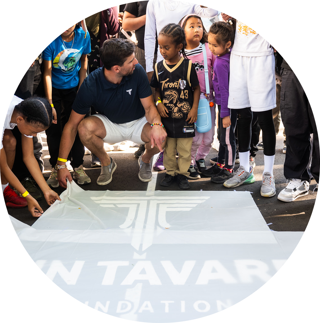 A group of children and a man gathered around a large banner or sheet on the ground with a logo and text for the 'TAVAR Foundation.' The man is kneeling and talking to a young girl in a Toronto Raptors jersey, while other children watch.