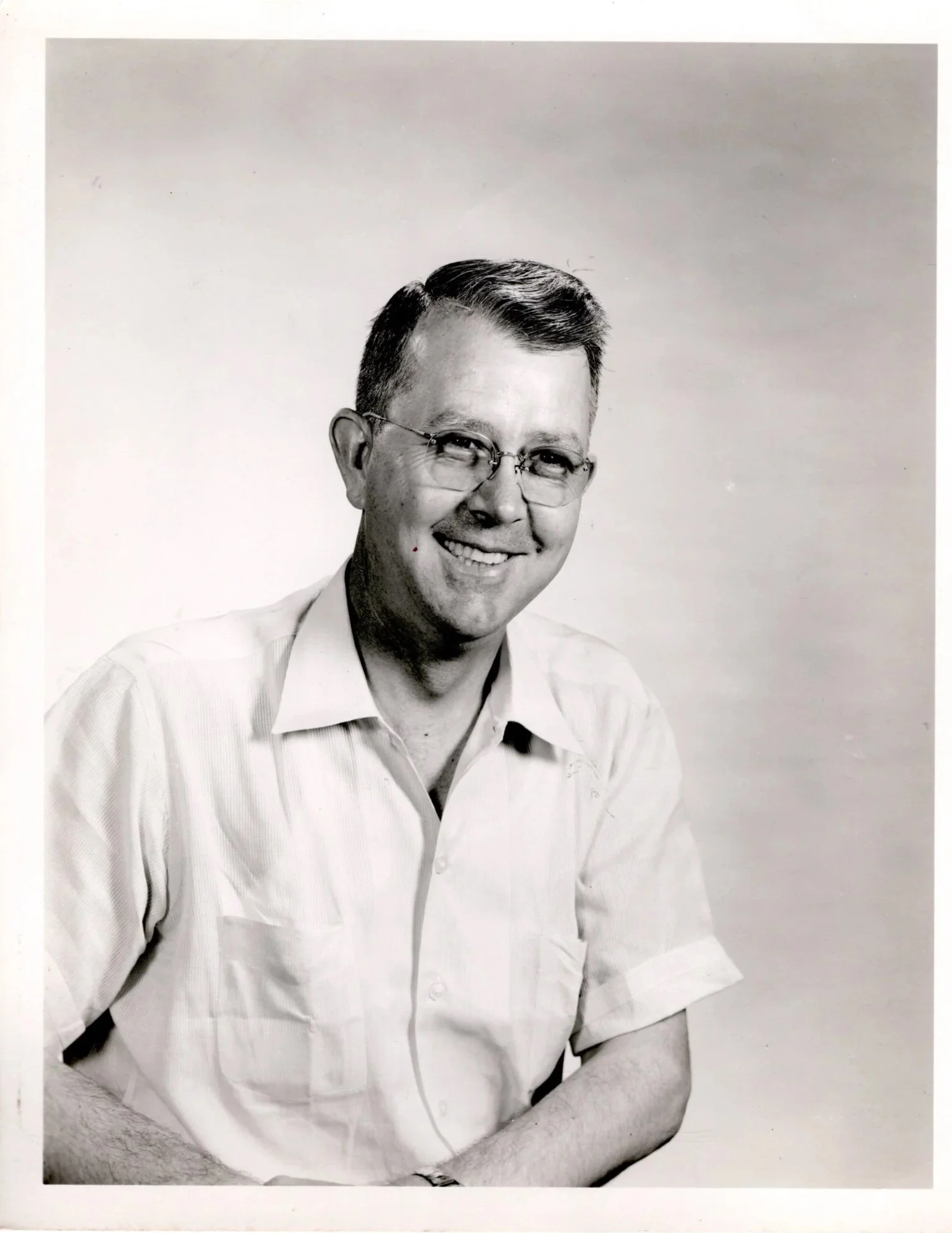 Black and white portrait of a man wearing glasses, smiling, and dressed in a collared shirt, with arms crossed.