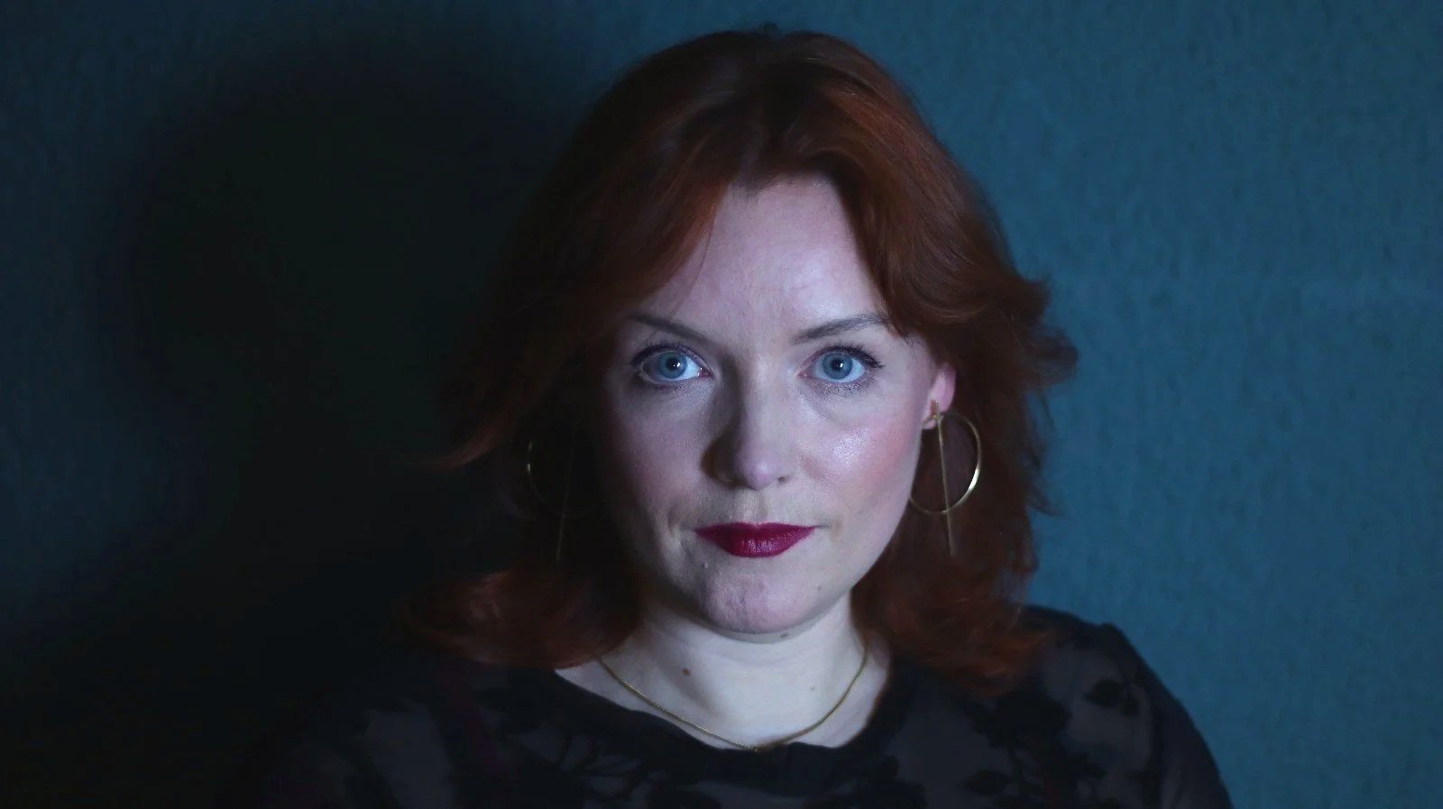A woman with red hair and blue eyes wearing hoop earrings, a gold necklace, and a black top with a pattern, posing against a dark background.