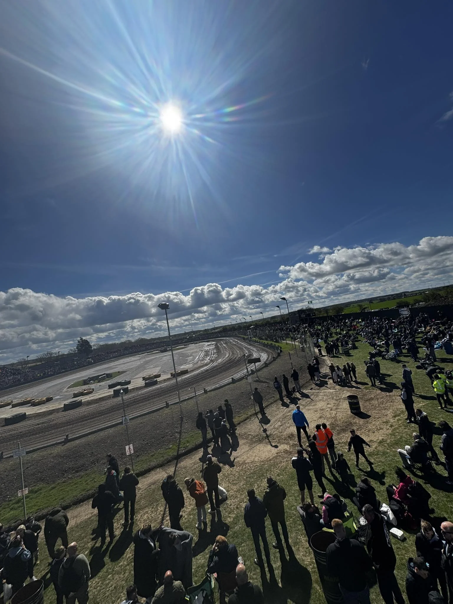 Crowd watching the speedway racing, under a bright sun with a blue sky and scattered clouds.