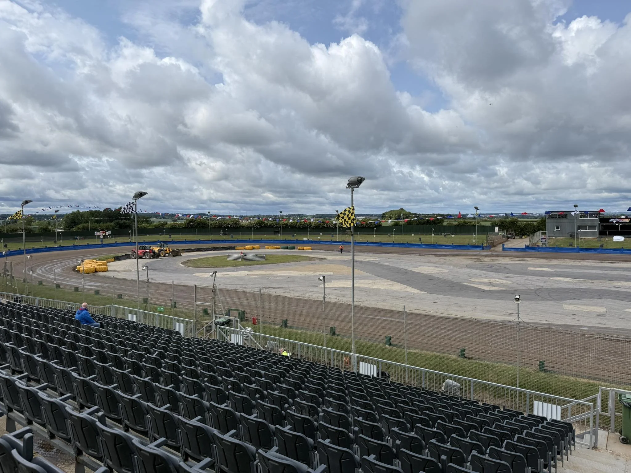 View of a race track with empty grandstands, a few people, and checkered flags, blue sky with clouds