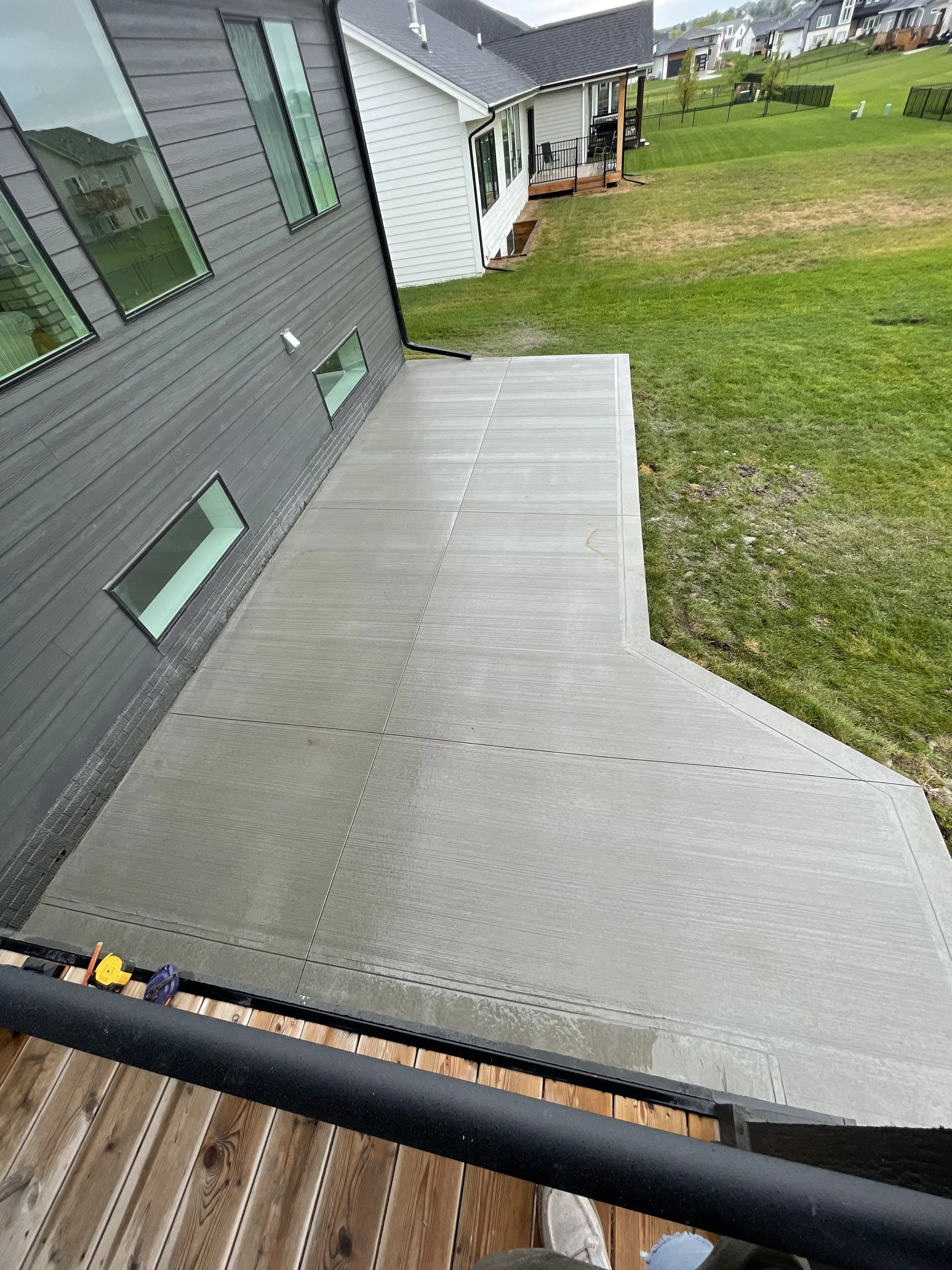 A view from a balcony showing a newly poured concrete patio, a grassy backyard, and neighboring houses with patios and fencing.