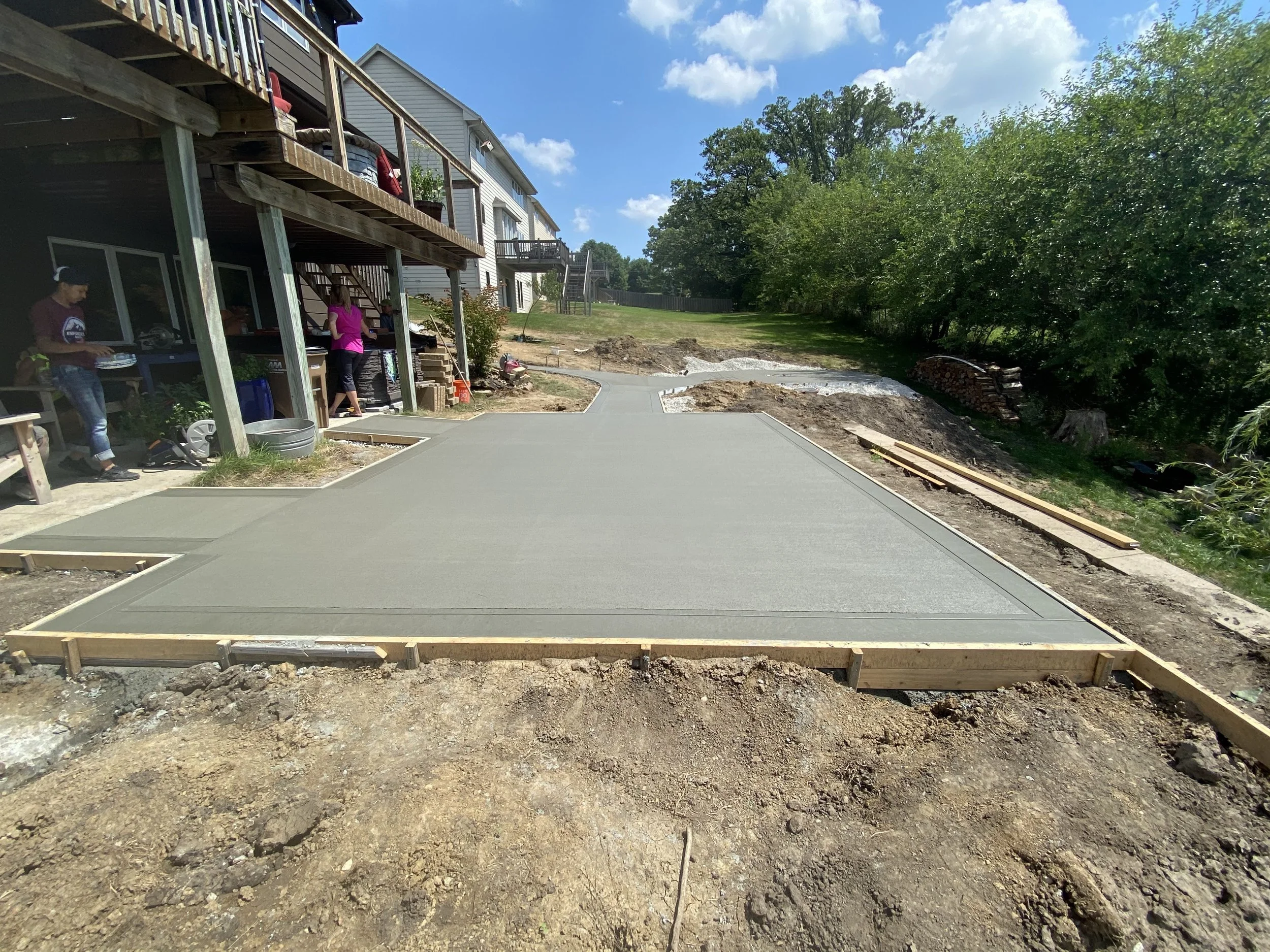 Freshly poured concrete driveway under construction in a residential backyard, with workers and construction materials visible.
