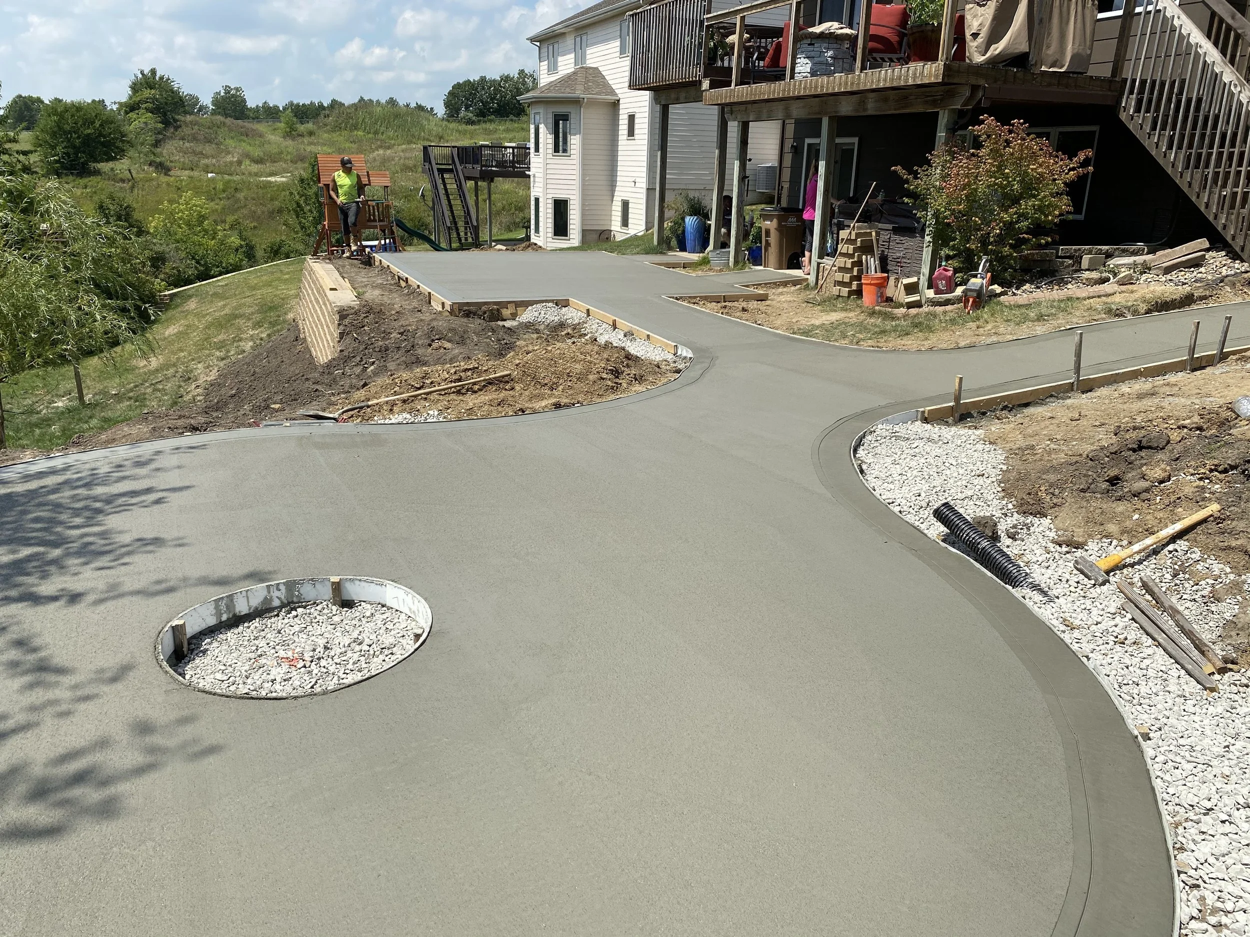 A newly poured concrete driveway curves around a residential property, with construction tools and materials visible on the ground.