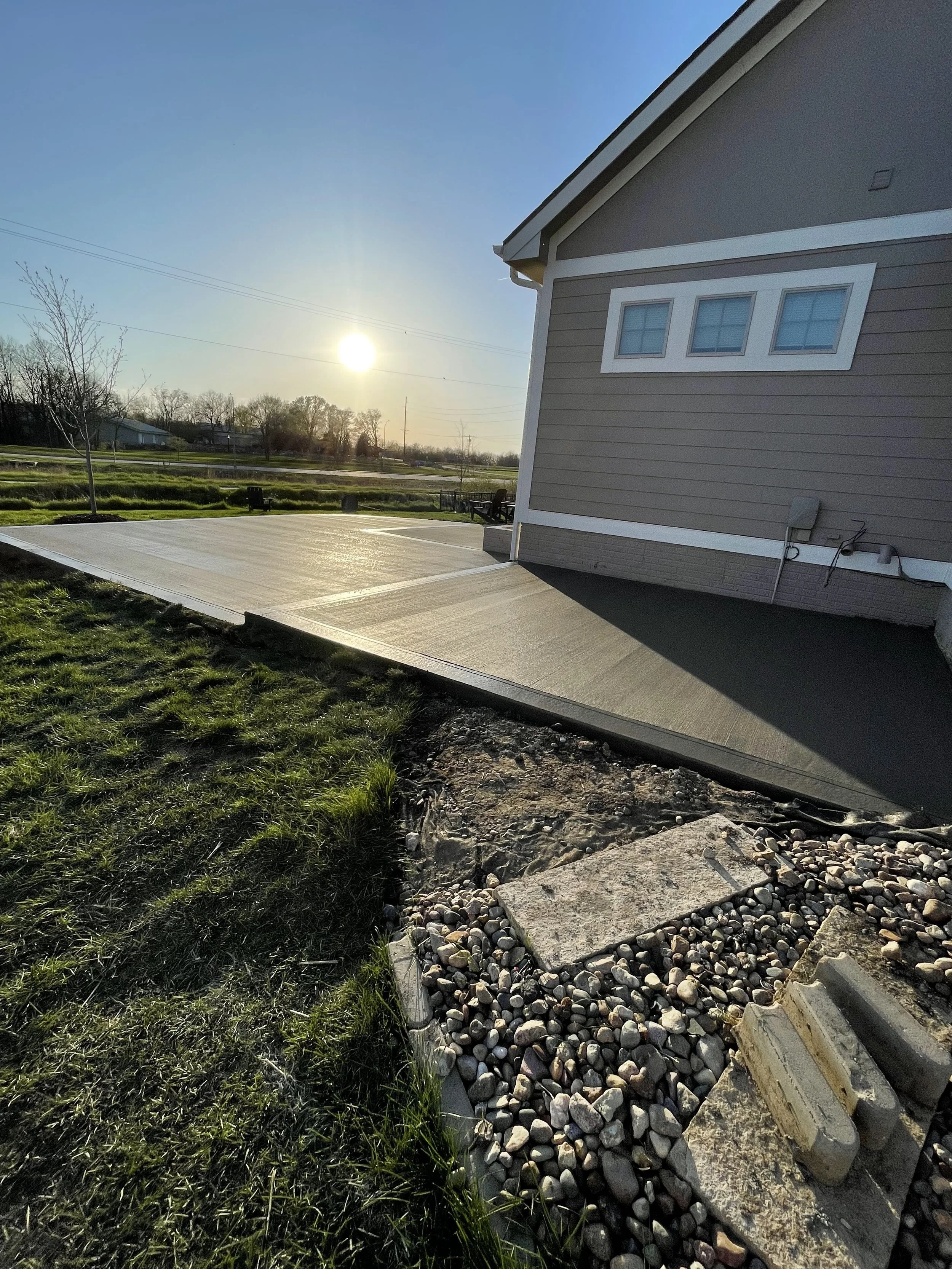 View of a freshly poured concrete patio adjacent to a house with beige siding and three small windows, in a neighborhood during sunset with trees and power lines in the background.