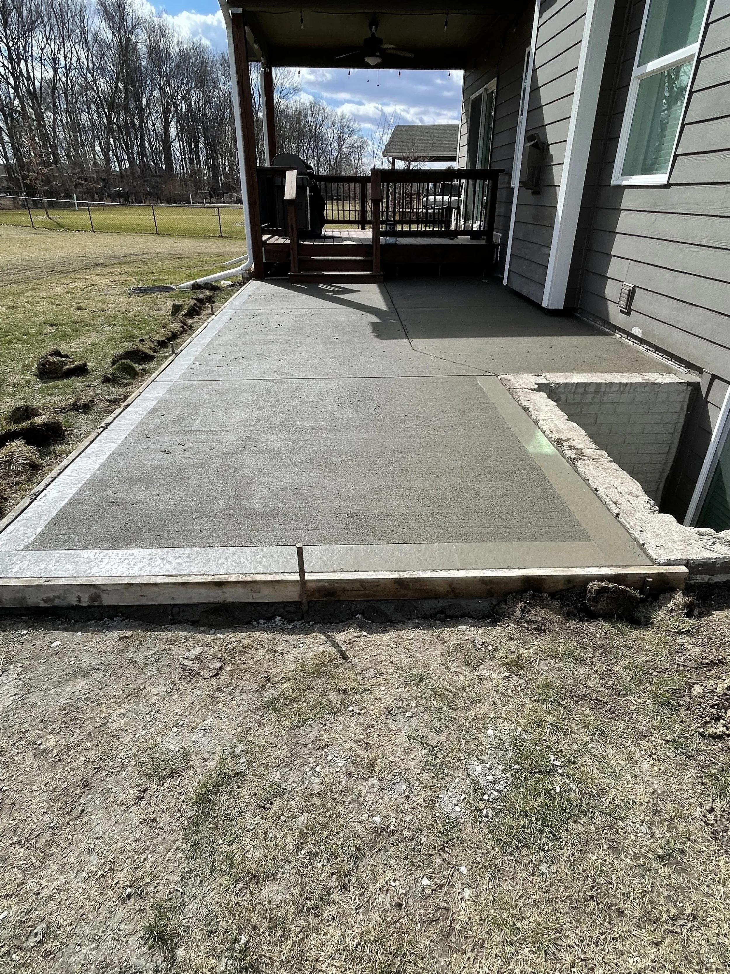 Concrete patio under construction at the back of a house, with a small porch and a wooden railing; the area is partially finished with some parts unpoured concrete and other parts smoothed, surrounded by a grassy yard and a chain-link fence in the background.