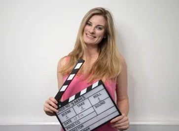 A smiling woman with long blonde hair holding a theater clapperboard, standing against a plain white background.