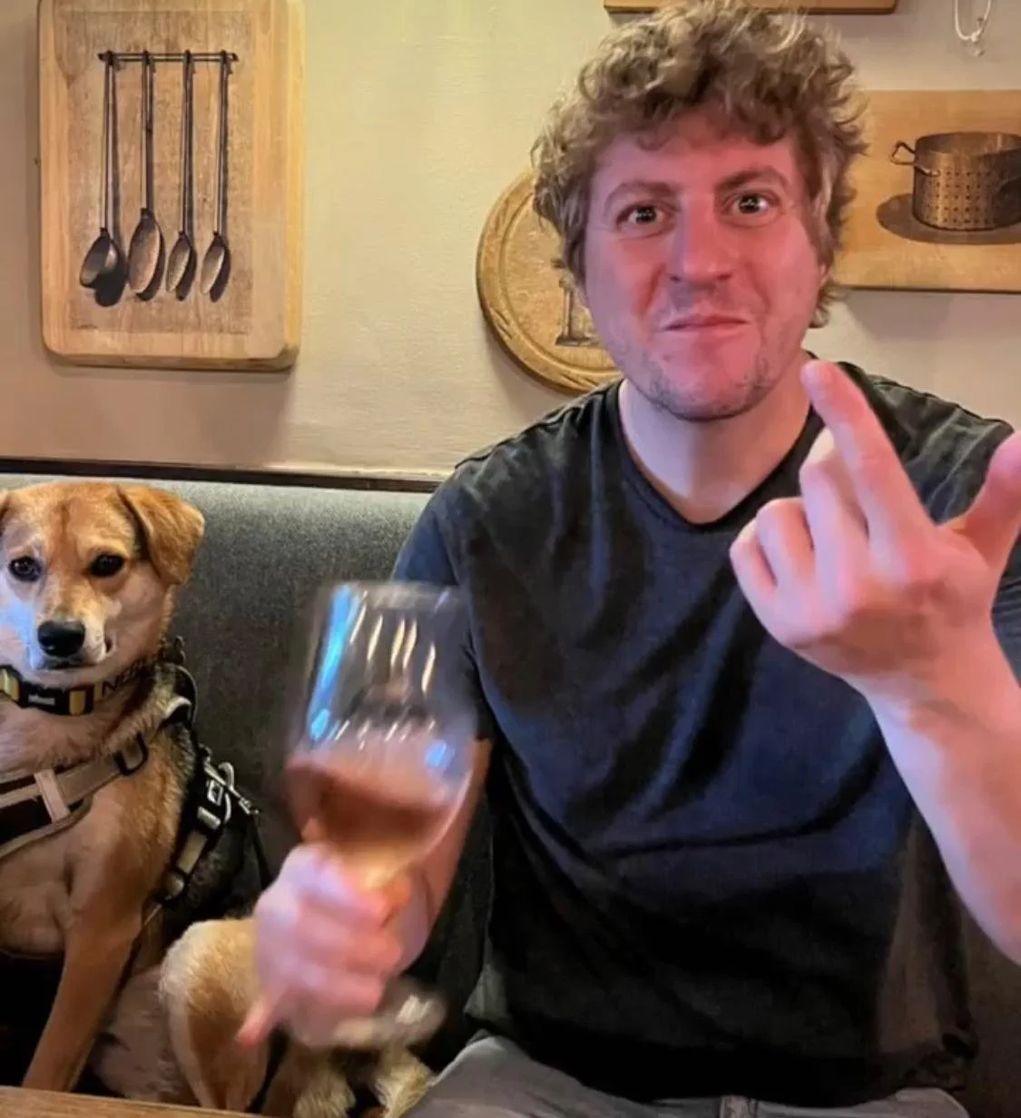 A man with curly hair making a gesture with his middle finger, holding a glass of beer, sitting on a couch with a dog beside him in a cozy room decorated with wooden kitchen utensils.