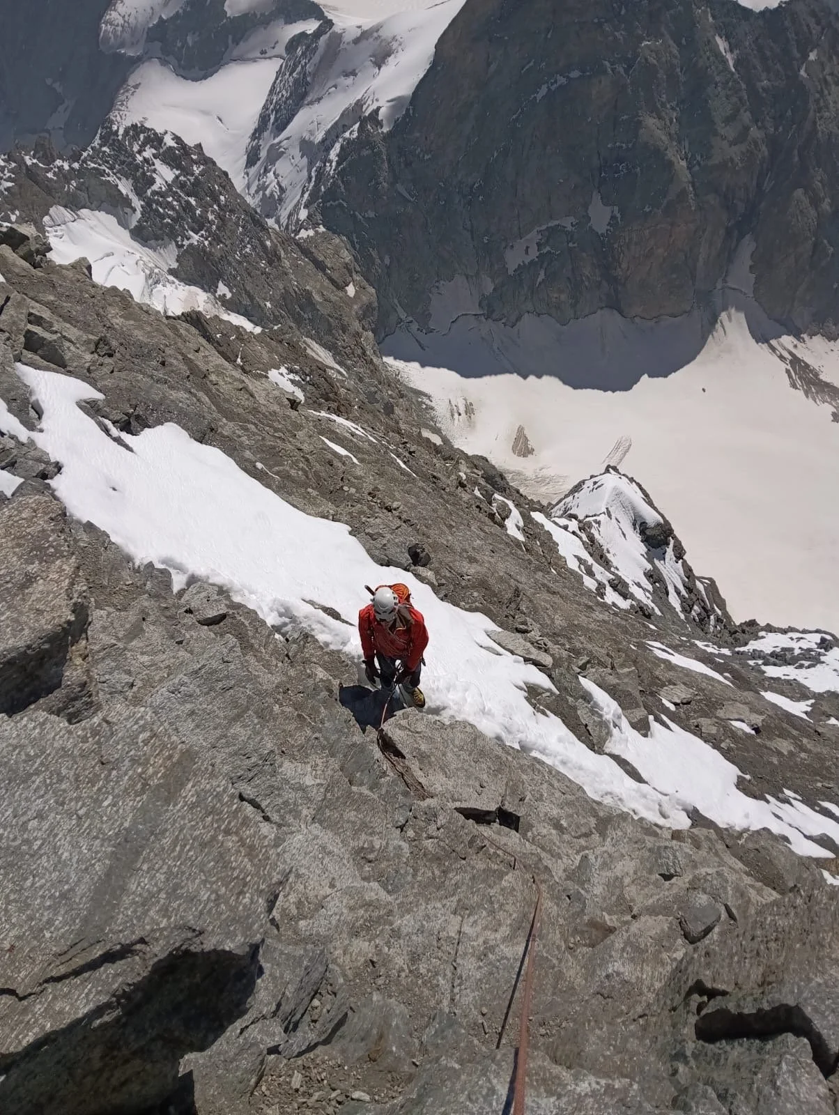 A climber in red gear ascending a steep rocky mountain slope with snow patches, using safety ropes and helmet, overlooking a glacier and mountain landscape.