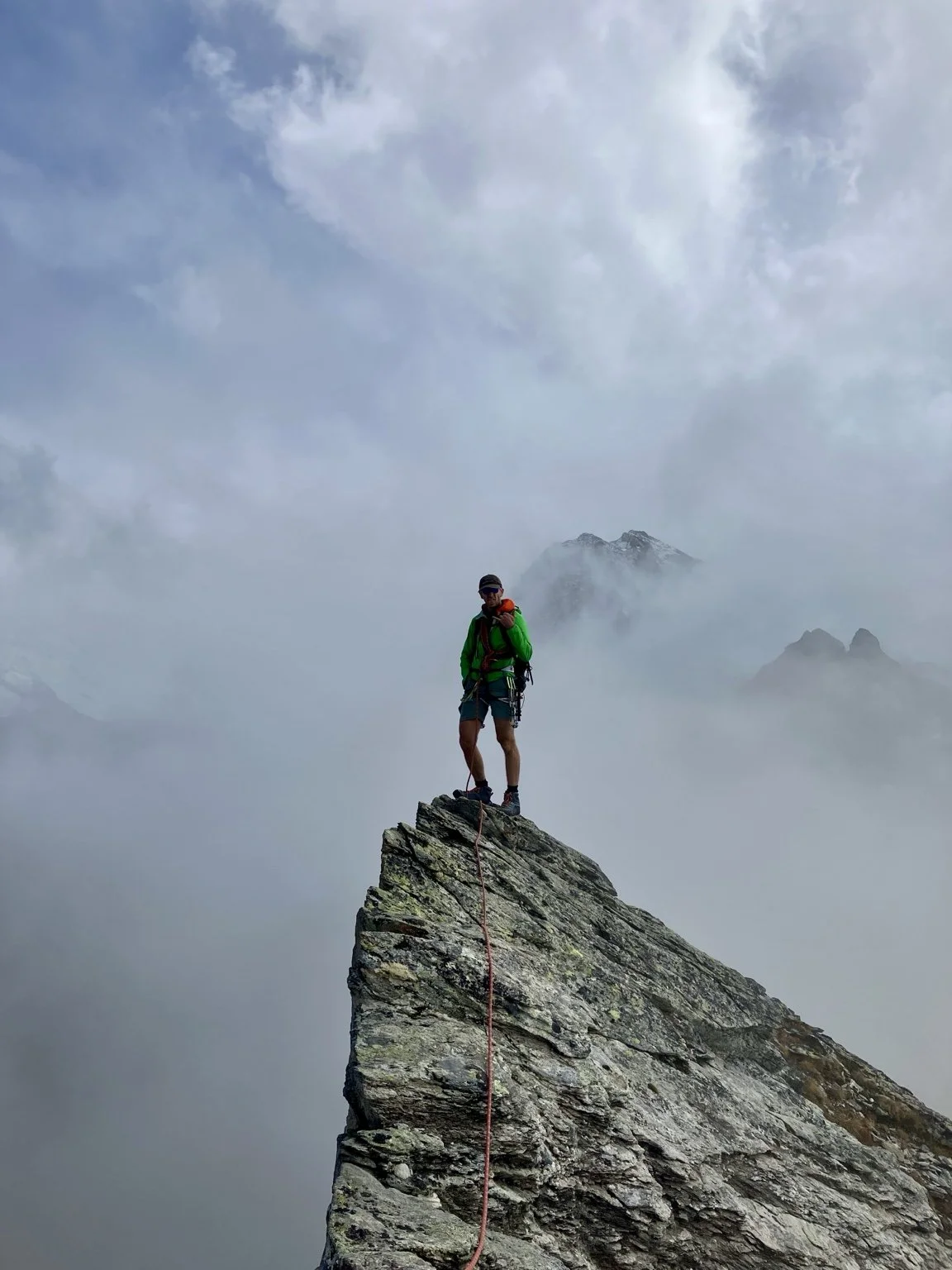 Une personne en randonnée en haut d'une falaise rocheuse, entourée de nuages et de montagnes, portant un sac à dos vert et des vêtements de randonnée.