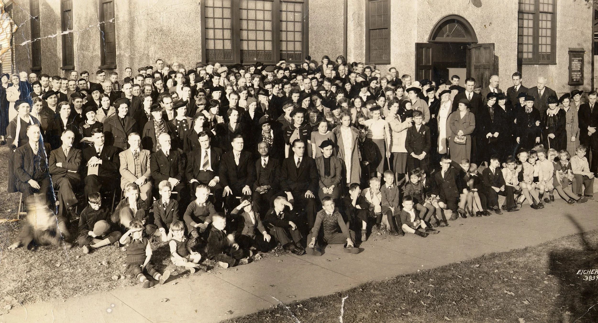 A large group of people, including children, teenagers, and adults, gathered outside a building for a group photo in black and white. The building has multiple windows and an open door.