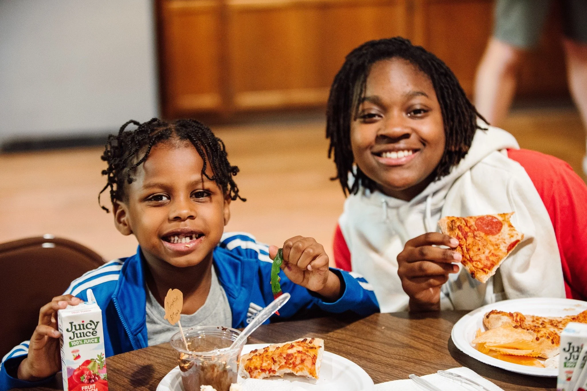 Two smiling children sitting at a table with pizza slices, juice boxes, and a small dessert, enjoying a meal together in an indoor setting.