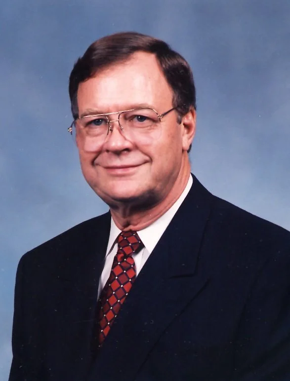 A man with glasses and dark hair, dressed in a dark suit and patterned tie, smiling against a blue background.