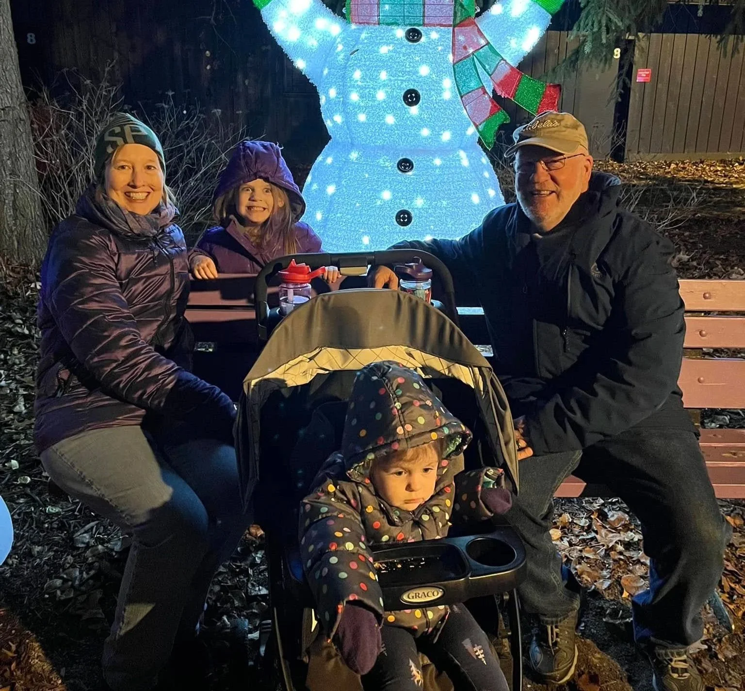 A family of four posing for a holiday photo outdoors at night, with a lit snowman decoration in the background. Two adults, a woman and a man, are smiling and kneeling beside a stroller that holds two children, one seated and one in a toddler seat. T