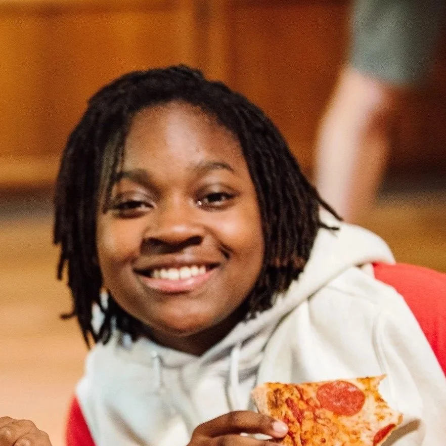 A young woman with dreadlocks smiling and holding a slice of pepperoni pizza.