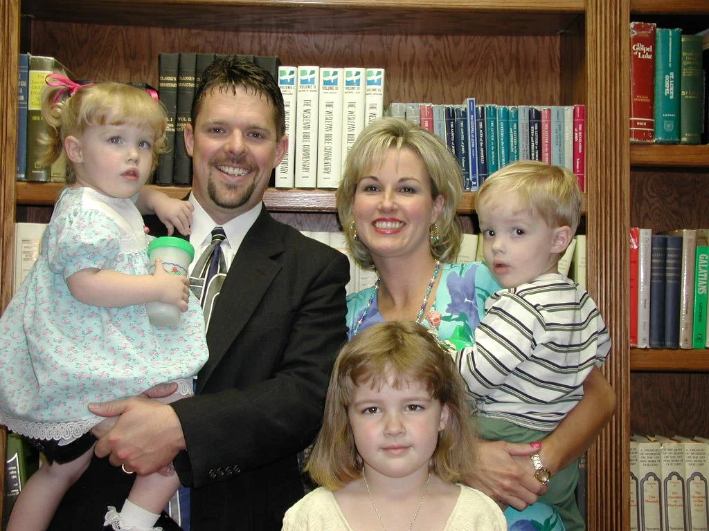 A family of five person poses for a photo in front of a bookshelf with old books. The family includes a man in a suit, a woman in a floral dress, and three children: a girl with red hair in a light-colored dress, a boy with blonde hair in a striped shirt, and a girl with light brown hair in a yellow shirt.