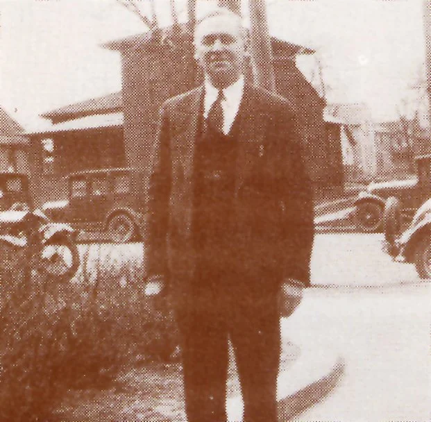 A black and white photo of a man in a suit standing outdoors near parked vintage cars, with a house and trees in the background.