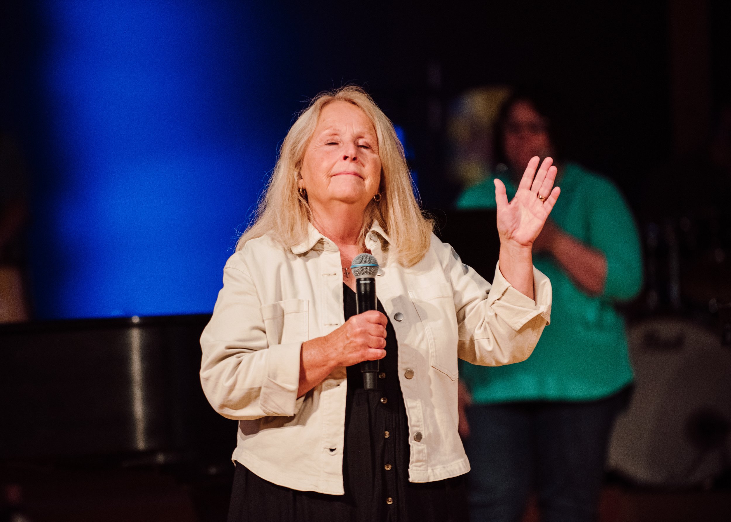 An older woman with blonde hair holding a microphone in her right hand and raising her left hand, appears to be speaking or performing on stage with a dark background and a person in green in the background.