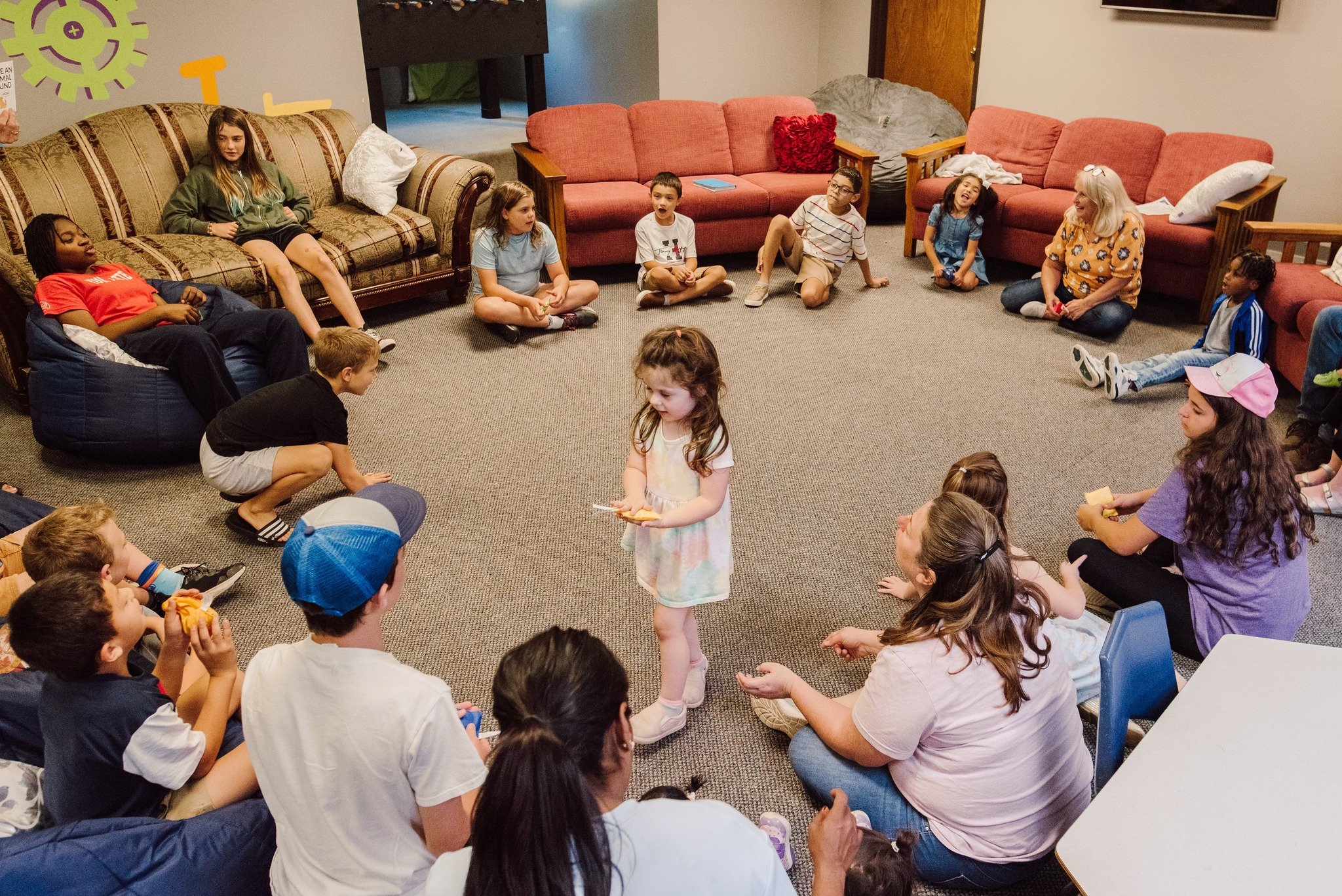 A group of children sitting in a circle on the floor in a cozy room, with one girl standing in the center holding a piece of paper, likely during a group activity or game.