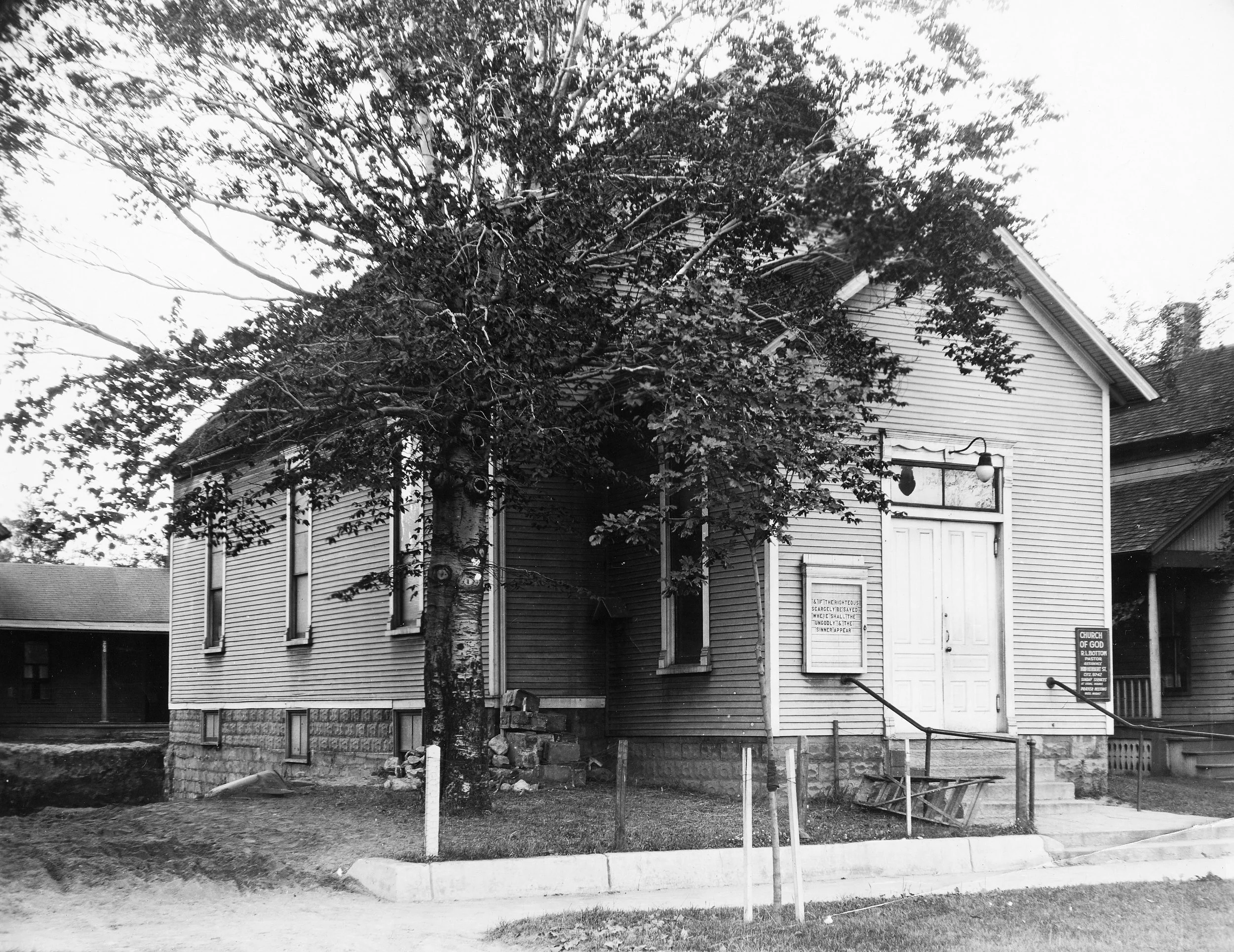 Black and white photo of a church building with white siding, a large tree in front, and a small sign on the wall. The church has a double door at the entrance, steps, and exterior lights. Adjacent houses are visible in the background.