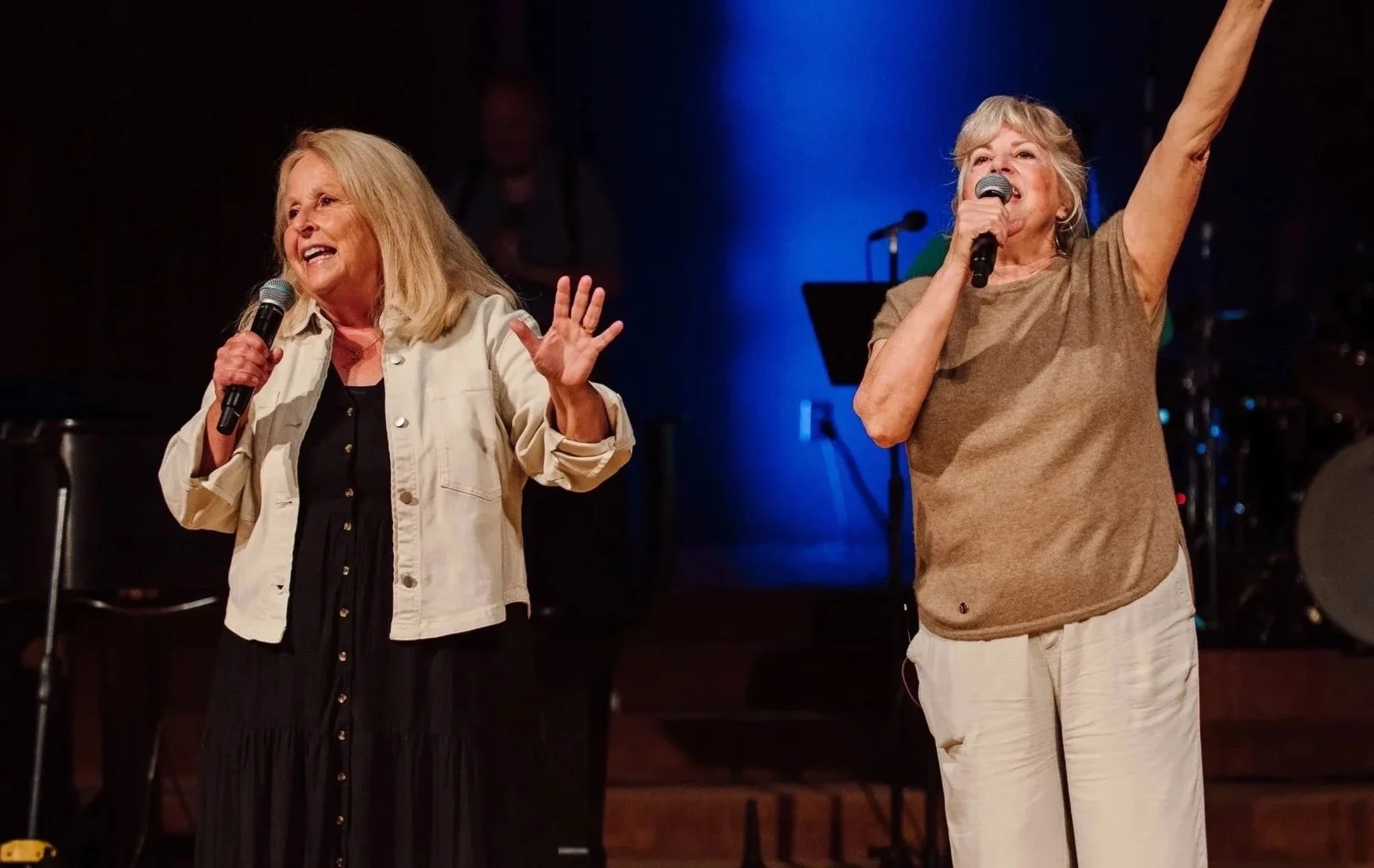 Two women singing on stage with microphones, one wearing a light jacket and black dress, the other in a brown shirt and light pants, raising her arm, with musical instruments in the background.