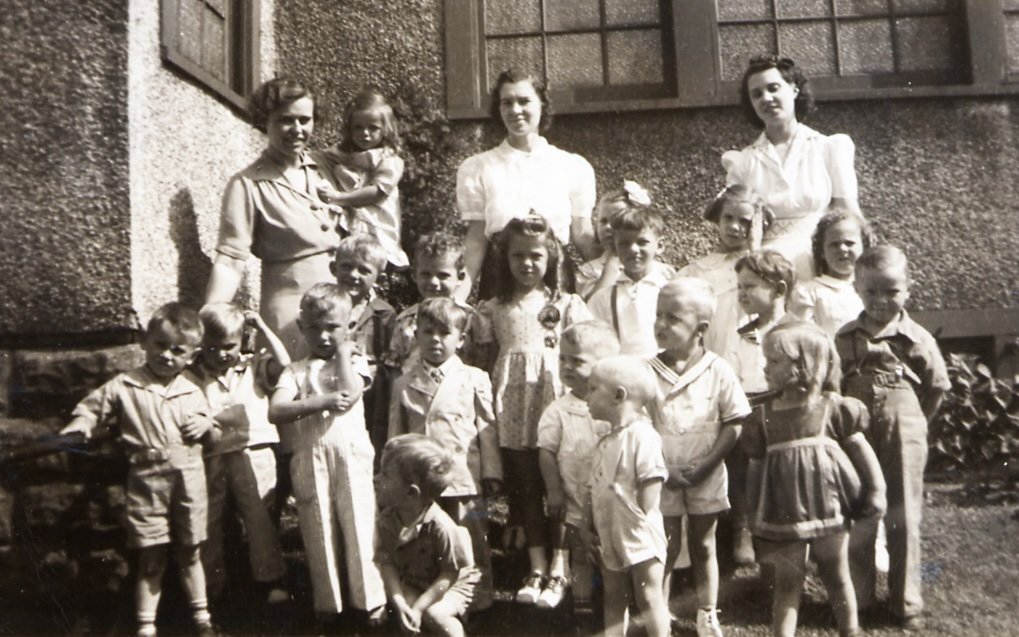 Black and white photo of a group of children and three women outside a building, likely from the mid-20th century.