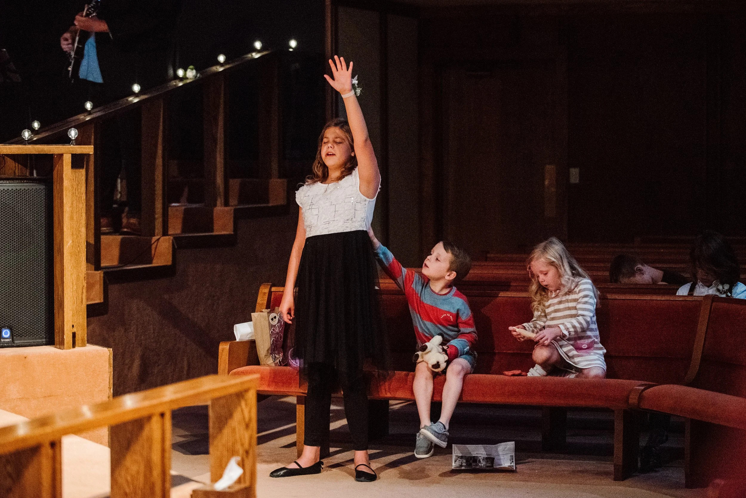 Children on a church pew, with a girl in a white top and black skirt singing with her eyes closed, a boy in a striped shirt with a toy, and a girl with a striped dress looking at her phone.