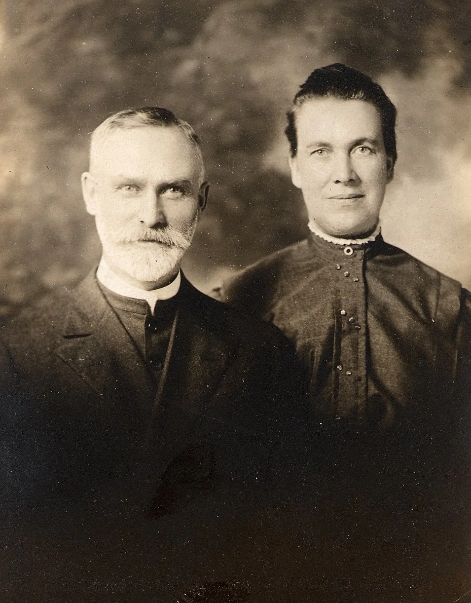 A sepia-toned vintage photograph of a man with a beard and mustache, and a woman with dark hair, both dressed in period clothing, posing against a blurred background.