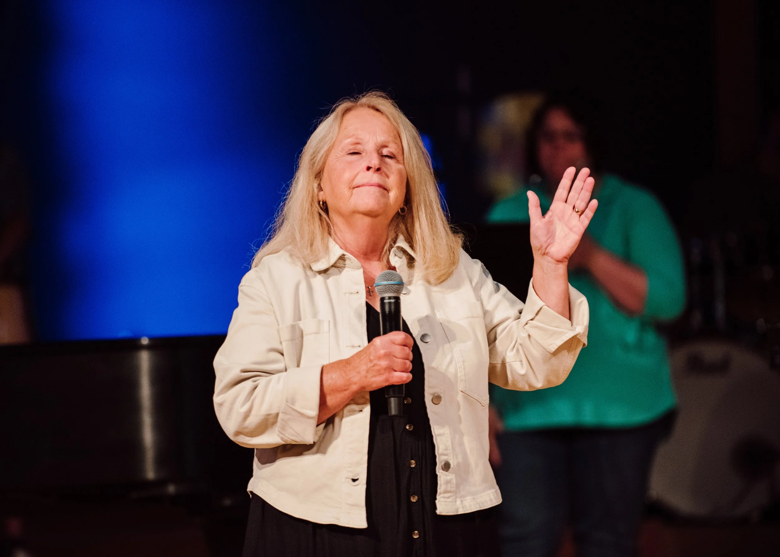 An older woman with blonde hair holding a microphone in her right hand, wearing a cream-colored jacket and a black top, with her left hand raised, at a live event or performance.