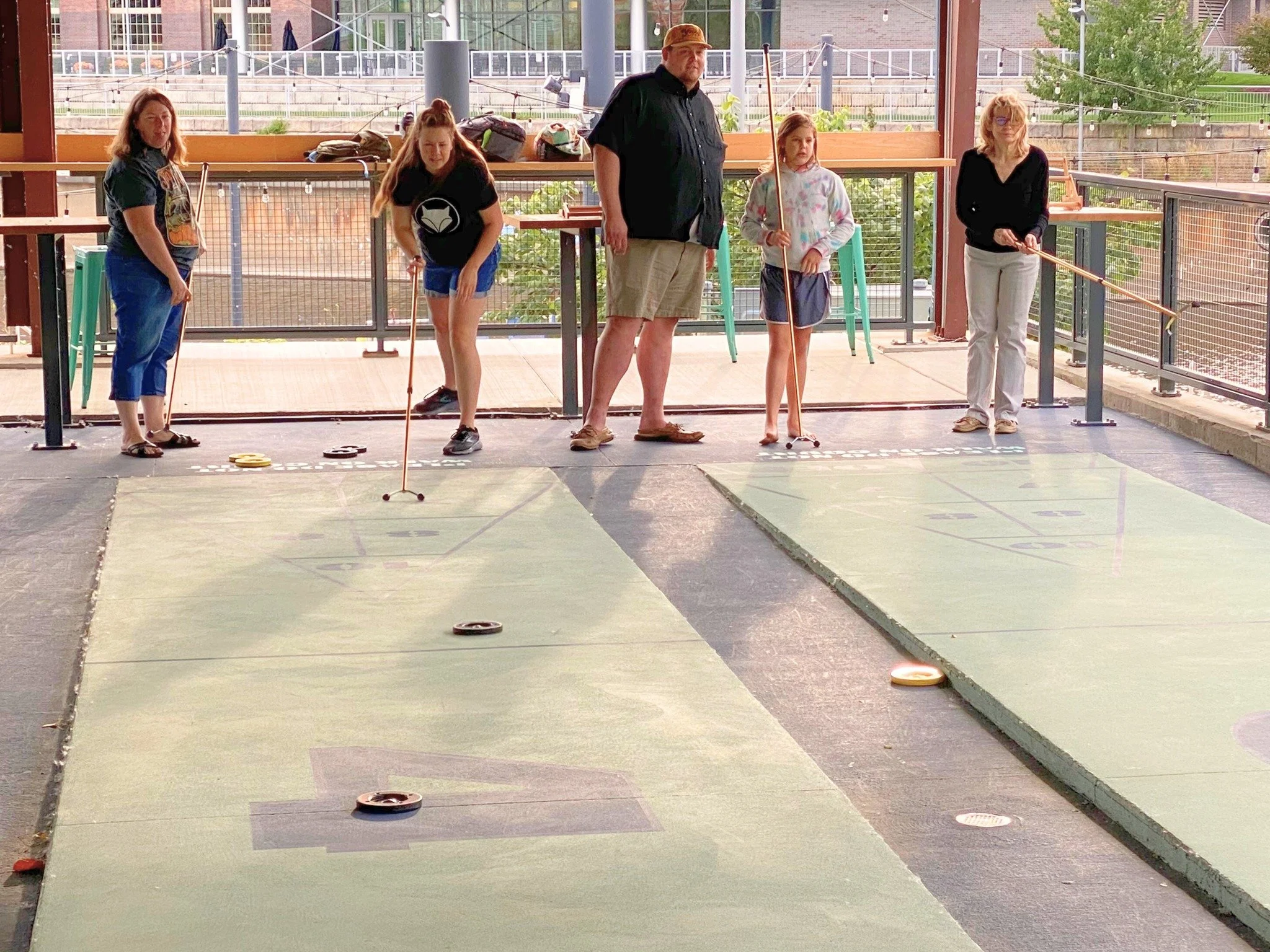 People playing indoor shuffleboard on two courts, with several participants holding cues and aiming at pucks on the green surface, overseen by a man in black shirt.
